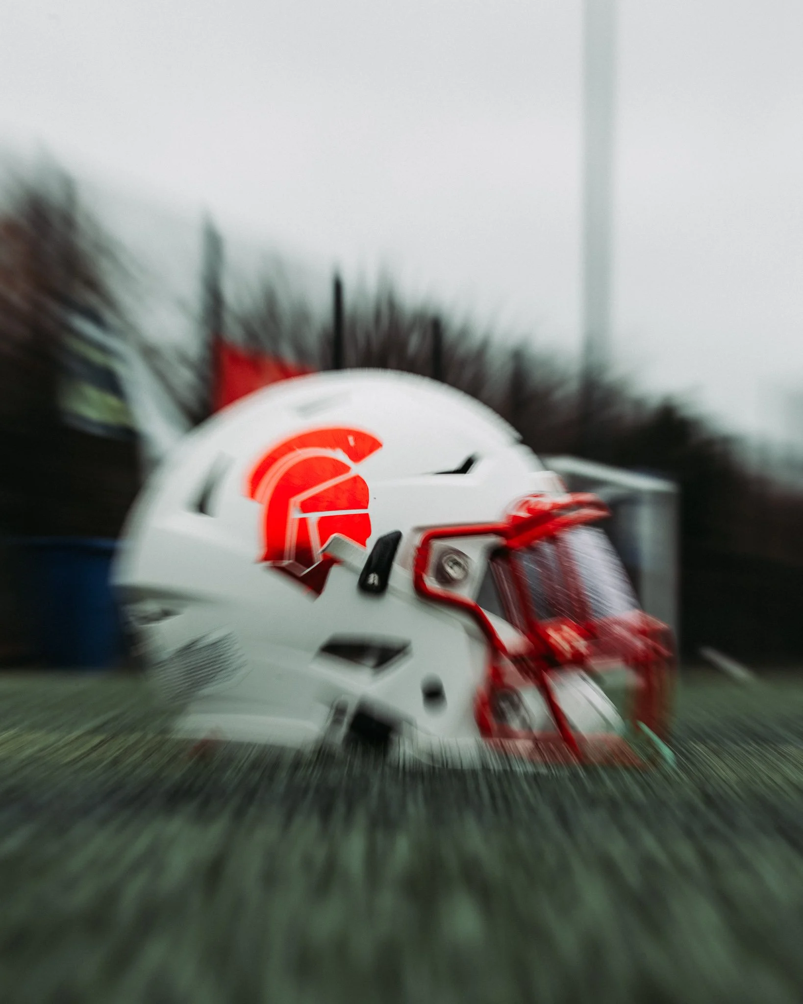 A white football helmet with a red and black Spartan logo and black faceguard, placed on the ground with a blurred background.