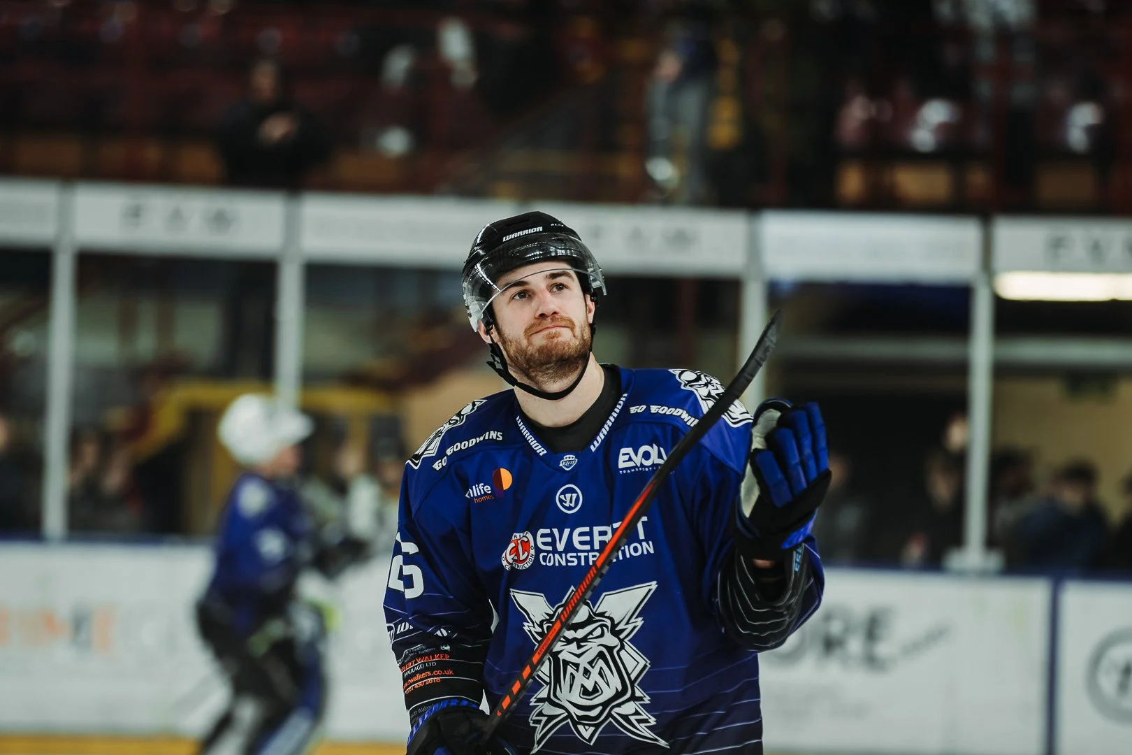 Ice hockey player in a blue jersey and black helmet looks upward on an ice rink, holding a hockey stick, with other players and spectators in the background.