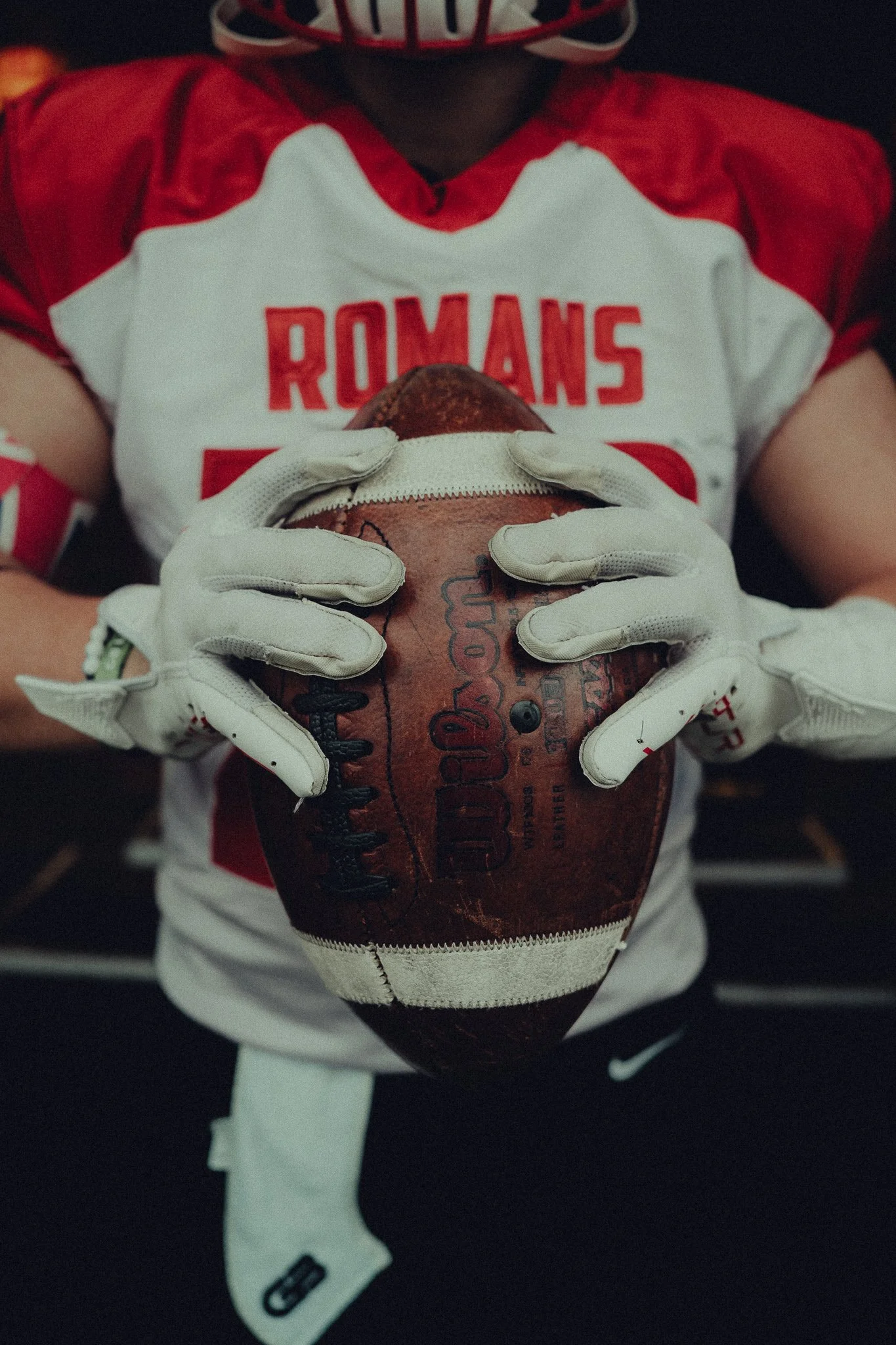 A football player in a red and white jersey holding a football with both hands. The jersey has 'ROMANS' written on it. The player is wearing white gloves and a red and white helmet and standing in a locker room.