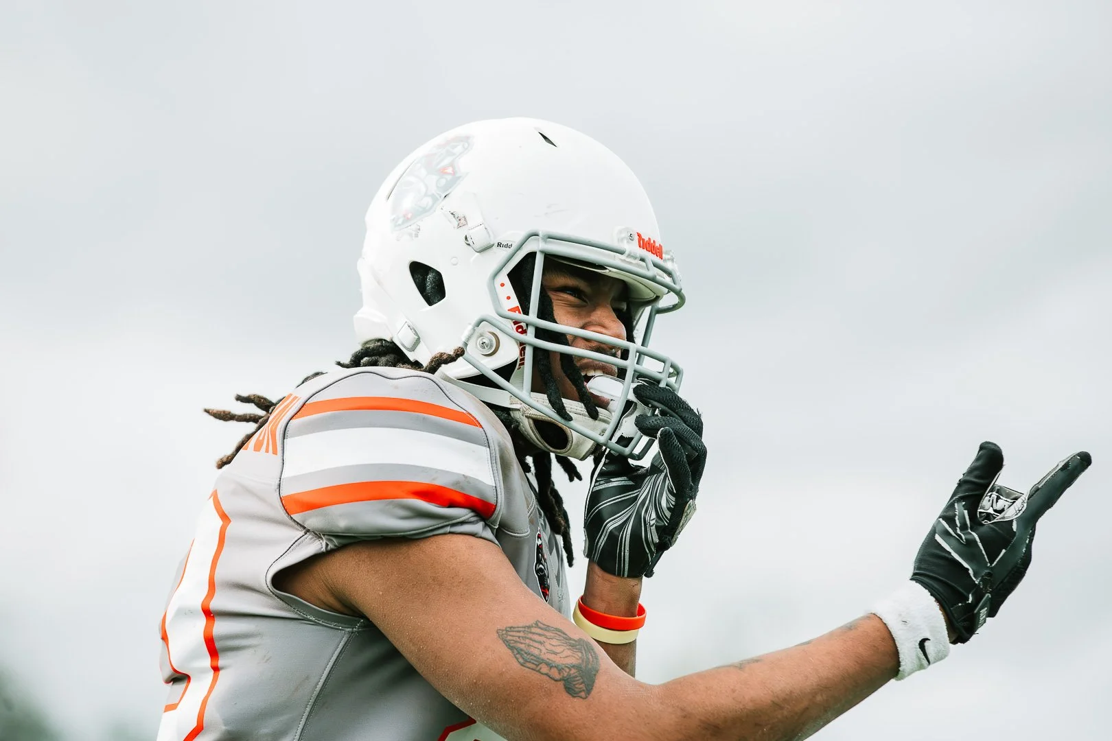 Football player wearing a white helmet and gray jersey with orange accents, smiling and pointing with two fingers during a game.