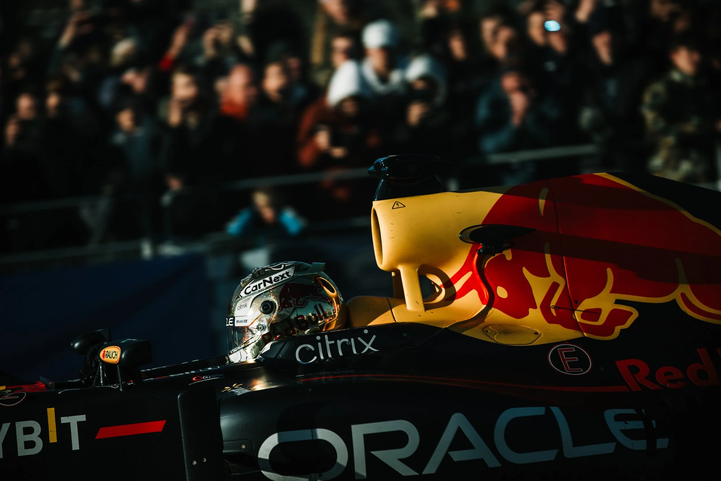 Close-up of a Red Bull Racing Formula 1 car at a race track with a driver in the cockpit, surrounded by a crowd of spectators in the background.