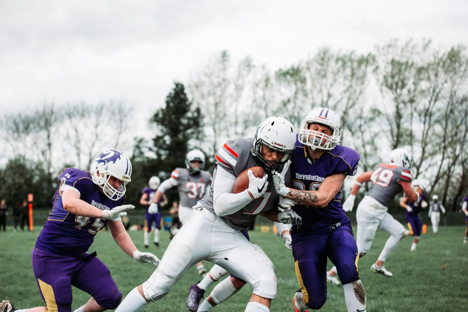 American football game with players in purple and gray uniforms, with a player running with the ball while being tackled by an opponent.