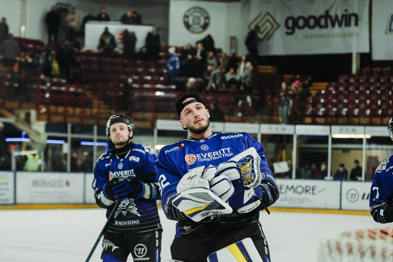 Ice hockey players in blue jerseys, one holding a hockey stick, on an ice rink during a game or practice, with spectators in the background.