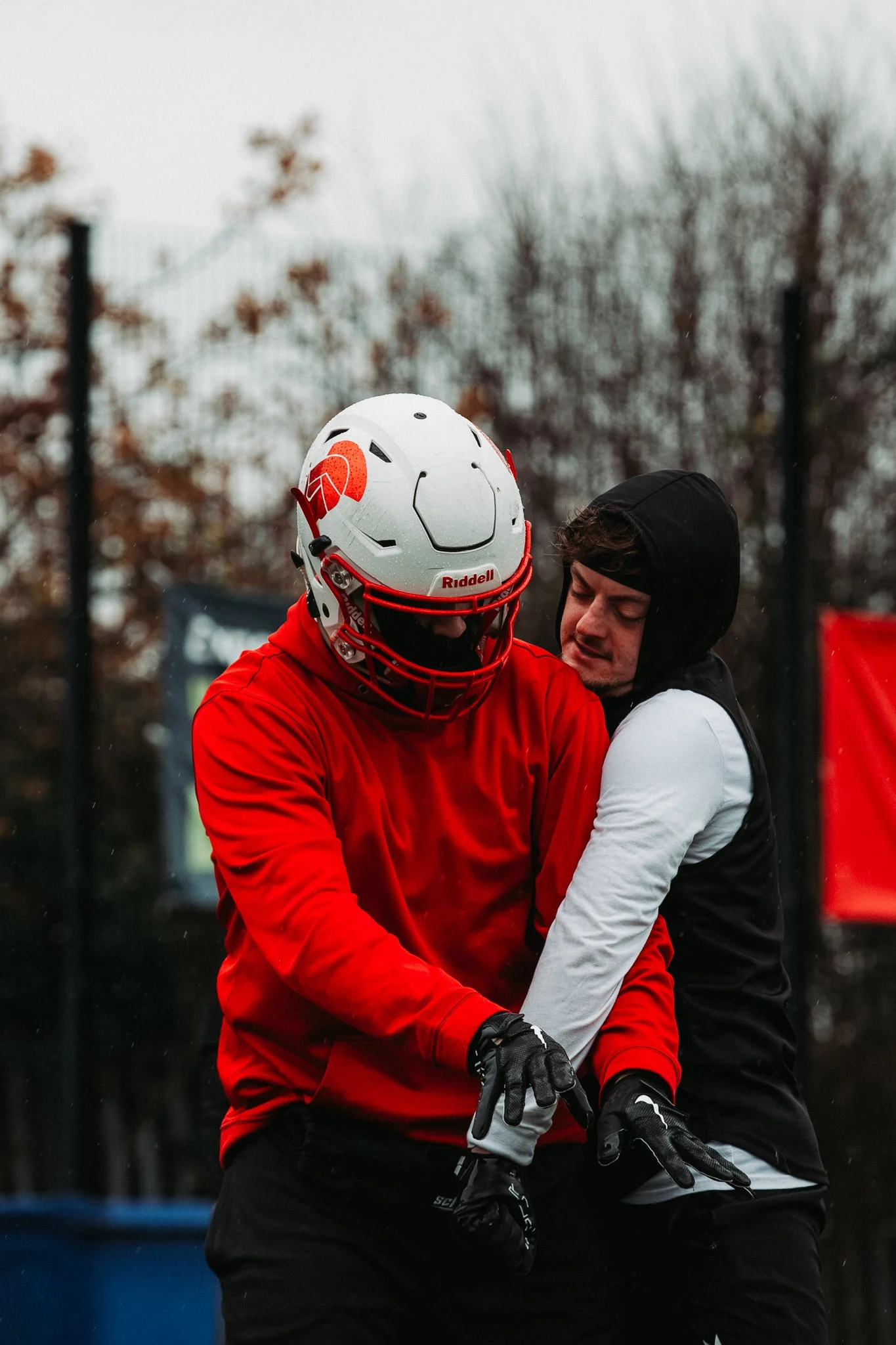 Two football players during a game on a rainy day, one in red and the other in black and white, with trees and a fence in the background.