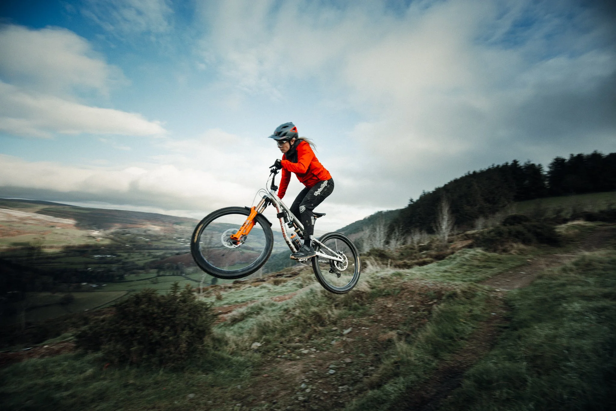 A person in a helmet and red jacket riding a mountain bike on a dirt trail in a hilly outdoor landscape.