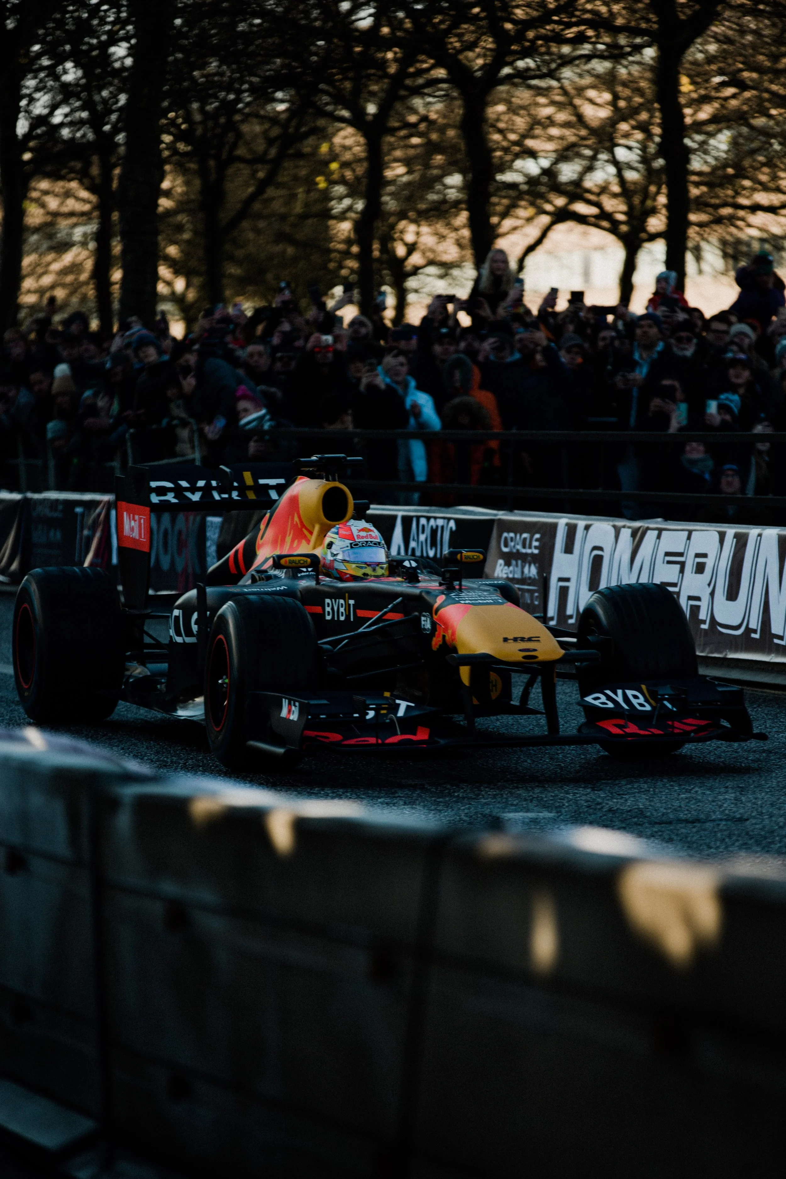 A Formula 1 race car with the Red Bull Racing team livery driving on a city street track during sunset, with a crowd of spectators watching behind barriers.
