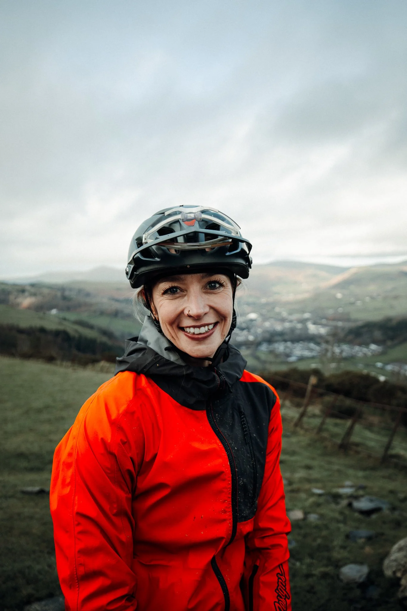 A smiling woman wearing a black bike helmet and a red and black jacket outdoors with a scenic hilly landscape and cloudy sky in the background.