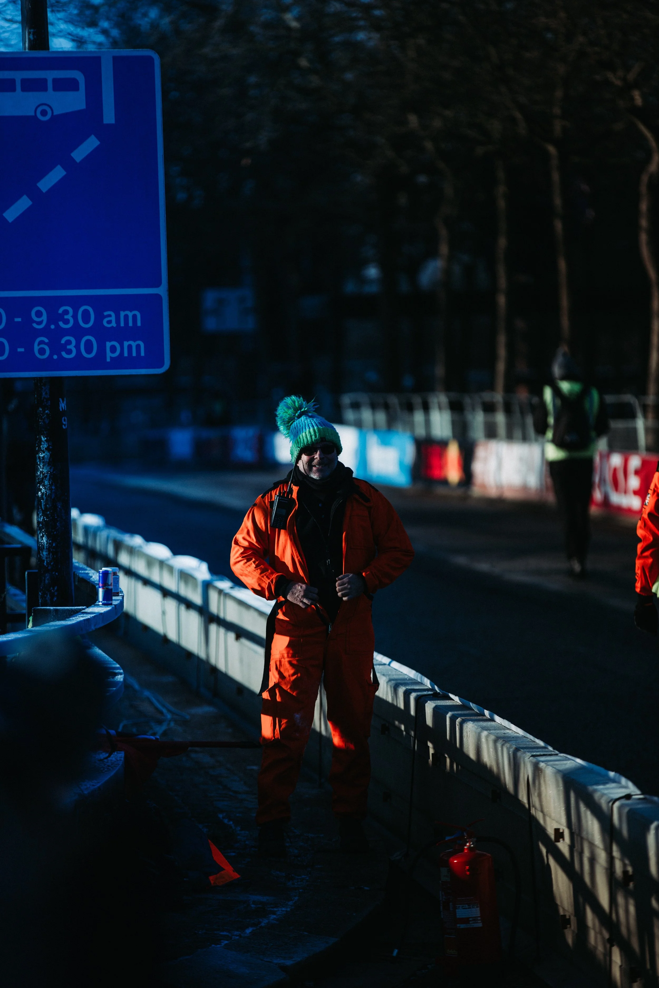 A person dressed in a bright orange jumpsuit and a colorful knit hat standing next to a metal barrier on a road at dawn or dusk, with other people in the background and a large blue sign indicating bus stop times.