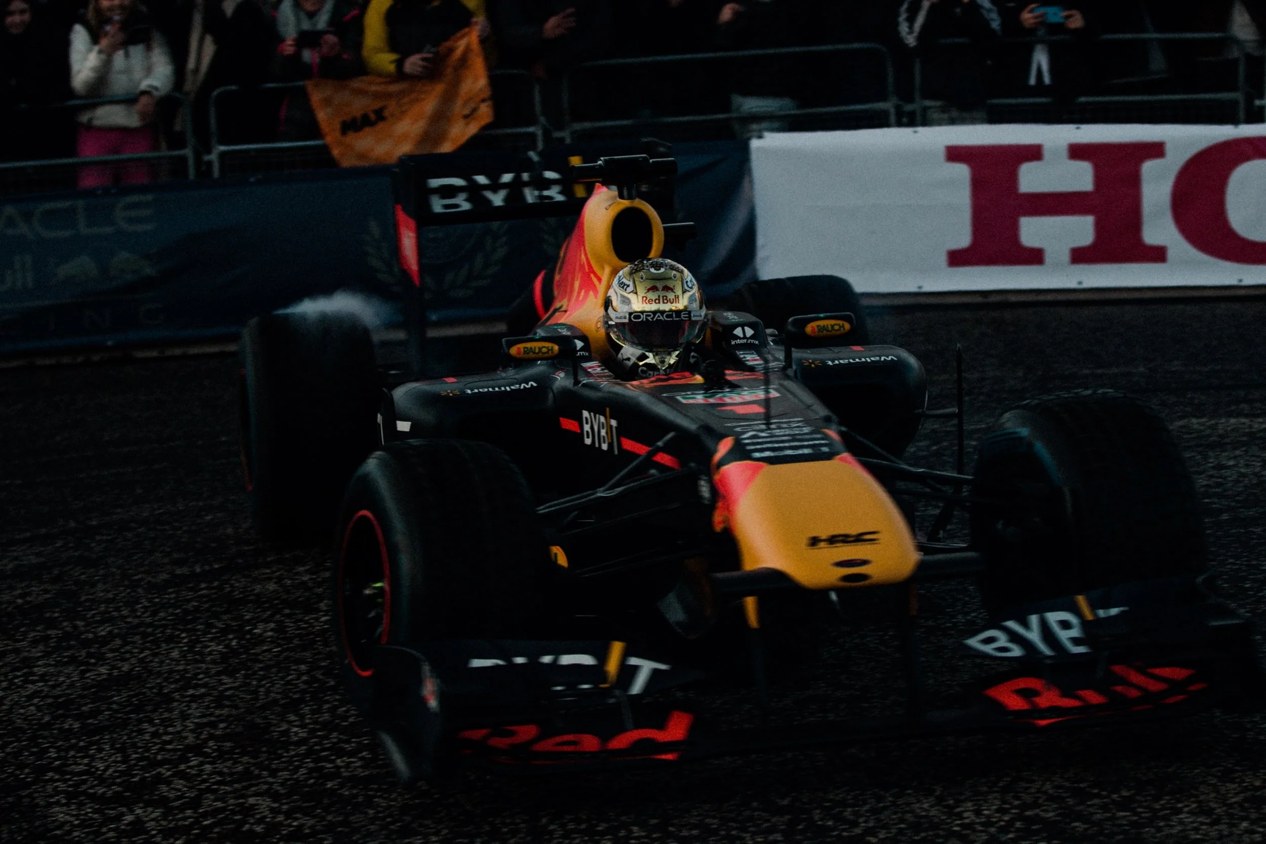 A Formula 1 race car with Black and yellow livery driven by a driver with a Red Bull helmet on a wet circuit, with spectators watching behind barrier barriers.