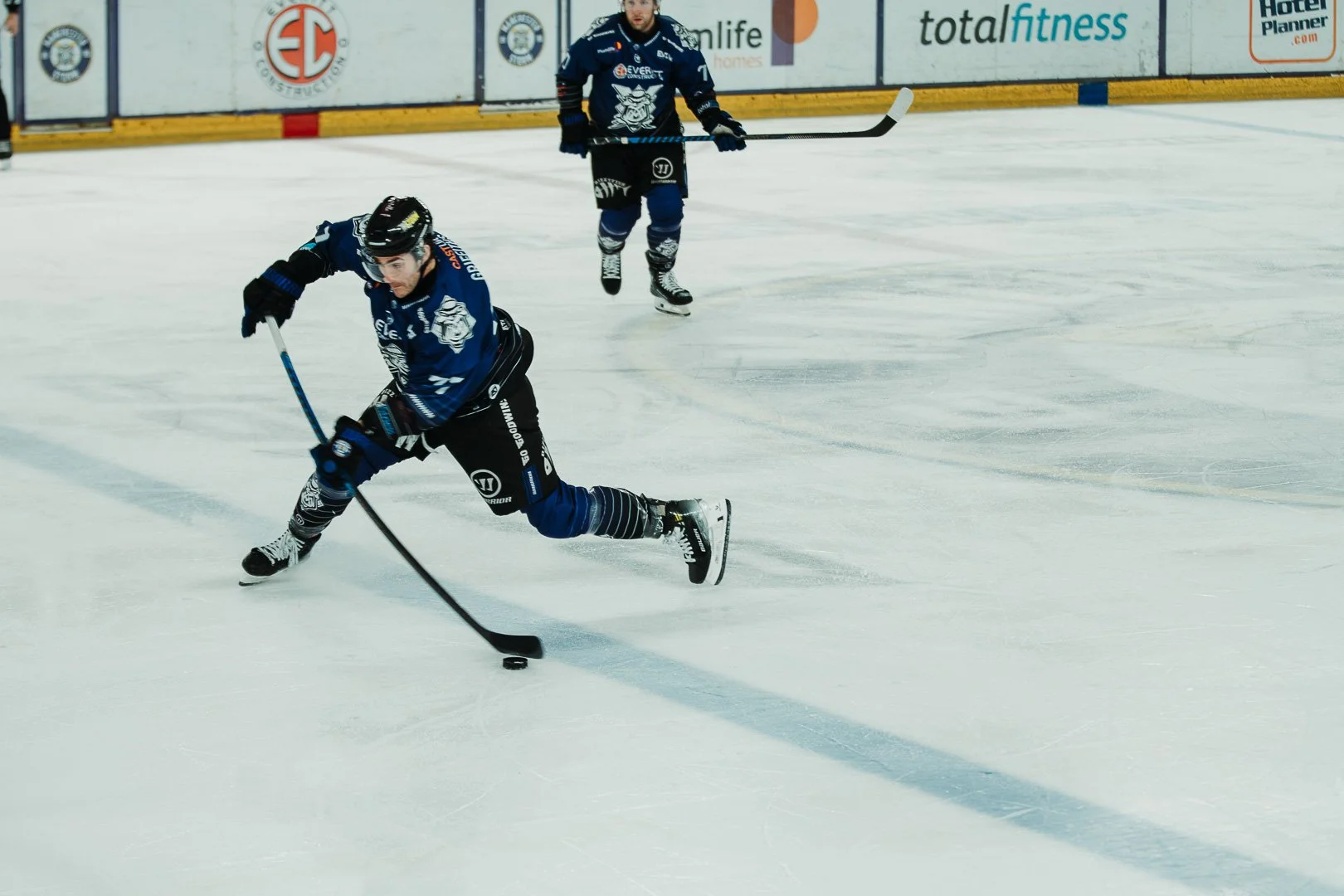 Two ice hockey players in blue jerseys playing on the ice rink, with one player in the foreground skating and controlling the puck, and another player in the background. The arena has advertisements along the boards.