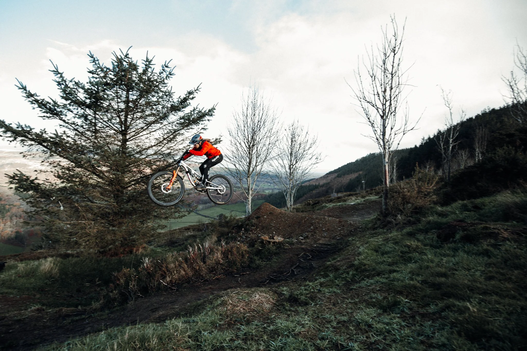 A mountain biker in a red jacket and helmet jumps off a dirt ramp on a trail in a hilly, wooded area during daytime.