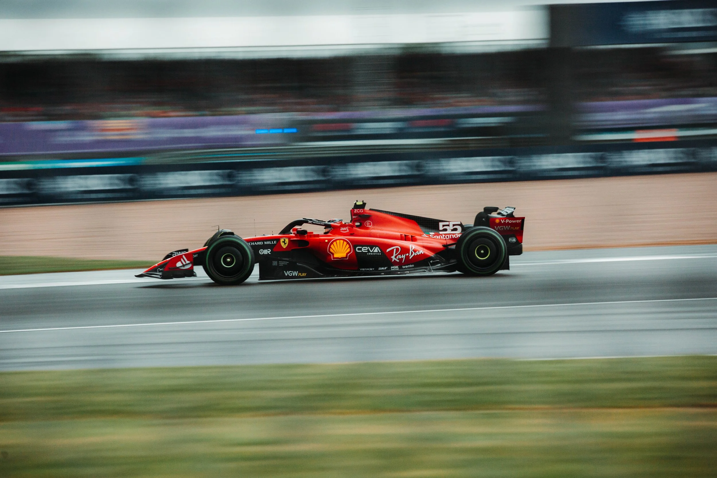 A red Formula 1 race car speeding on a race track, with sponsorship logos including Shell, Ray-Ban, Santander, and Ferrari, and a blurred background indicating motion.