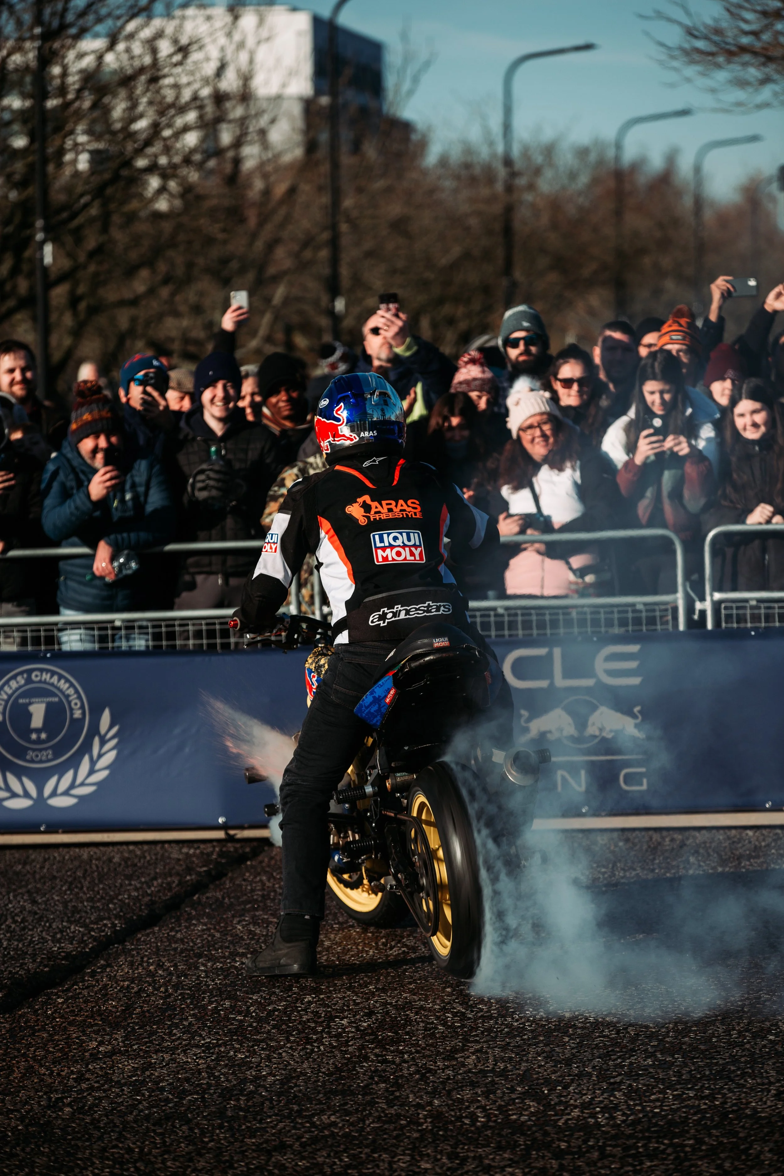 Motorsport event with a rider on a motorcycle doing a burnout, crowd of spectators taking photos in the background.