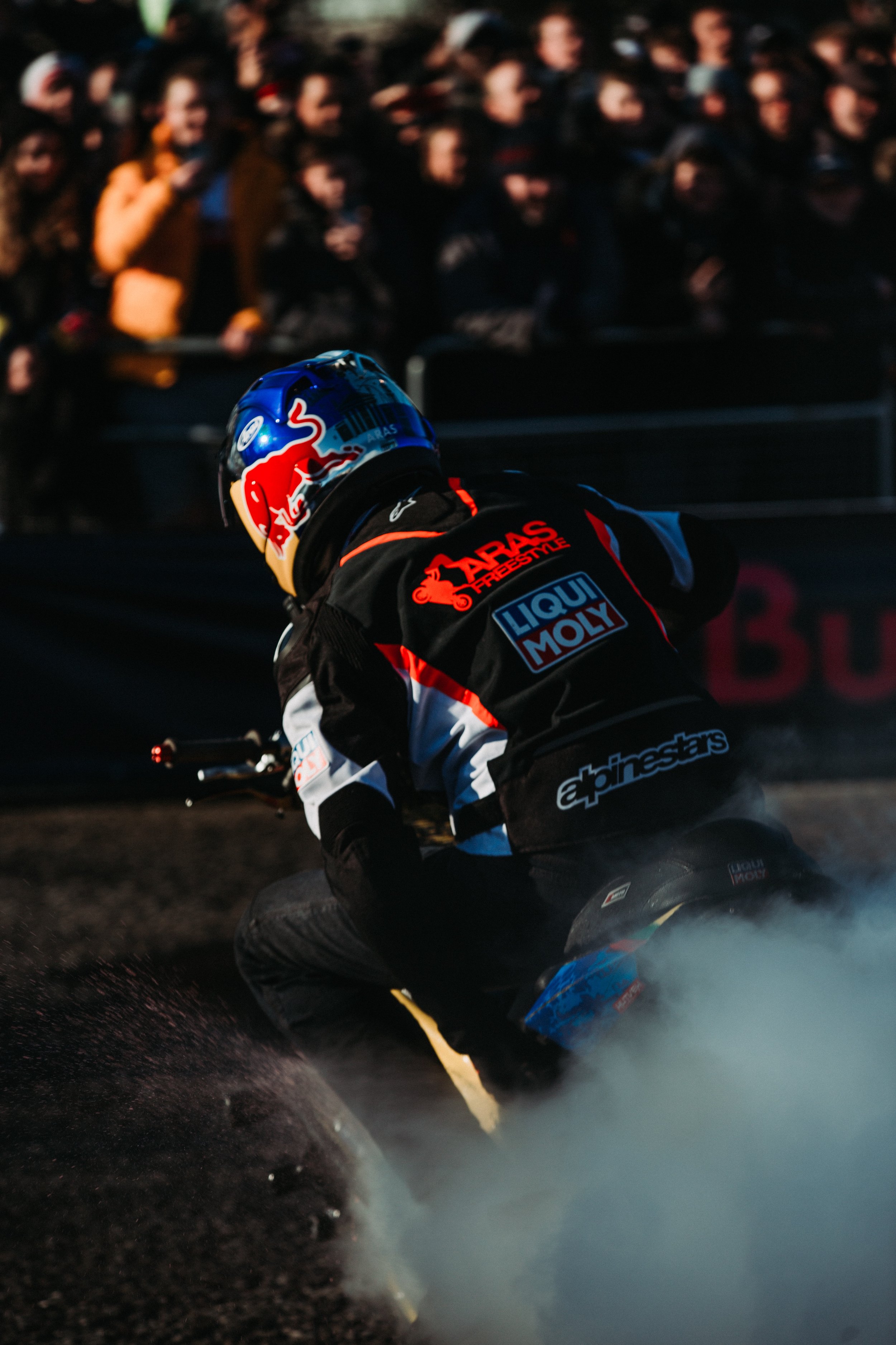 Motorsport rider performing a burnout on a dirt track with a crowd watching in the background.