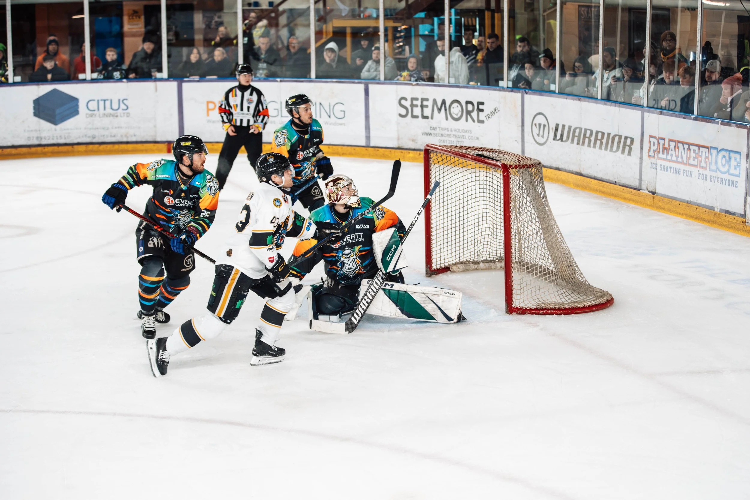 Hockey players and a goalie in front of the net during a game, with spectators watching from behind the glass.