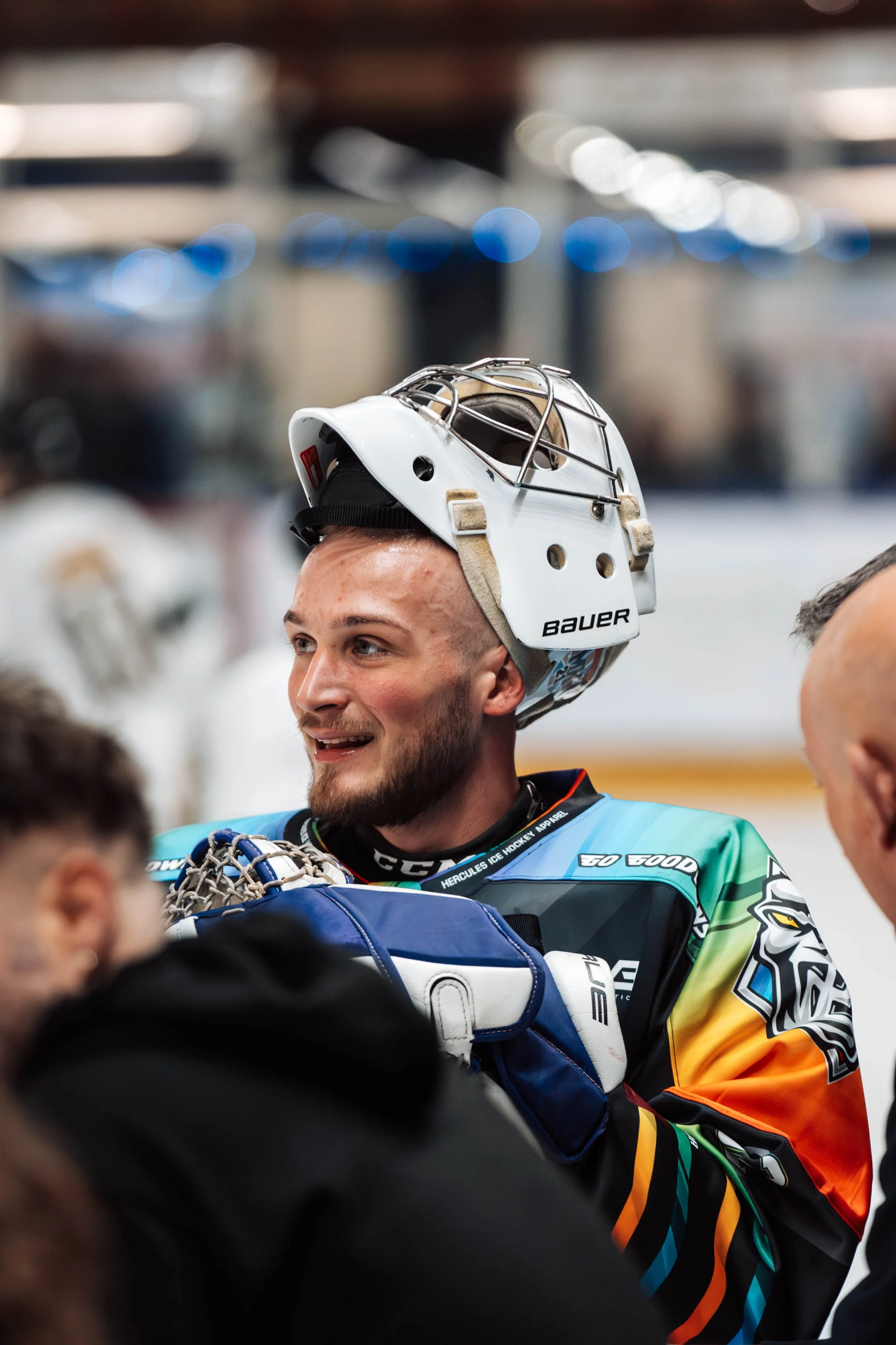 A young male ice hockey player with a beard smiling and wearing a colorful jersey, holding a goalie stick, and wearing a goalie helmet with a cage, in a rink setting.