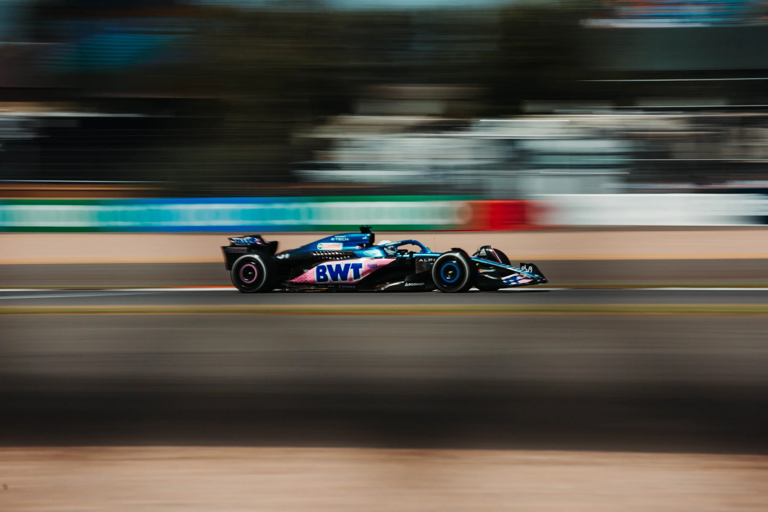 A Formula 1 race car speeding on a race track, with a blurred background indicating high speed. The car features the BWT logo and is primarily black with blue and pink accents.