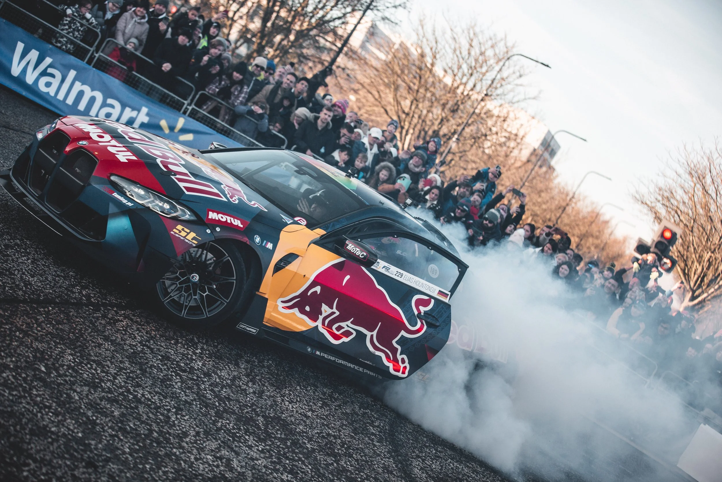 A racing car with Red Bull branding performs a burnout on a street, creating smoke, while a crowd of spectators watches behind barriers during daylight.
