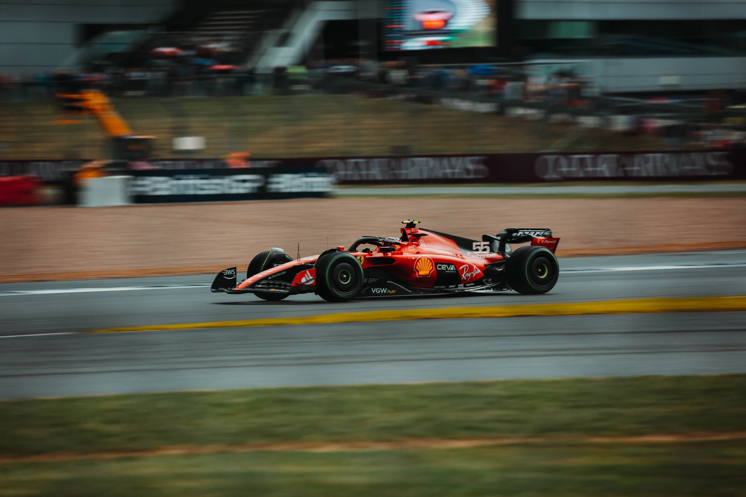 A red Formula 1 race car speeding on a race track with sponsorship logos, including Shell and VA, during a race. The background shows blurred spectators and trackside infrastructure.