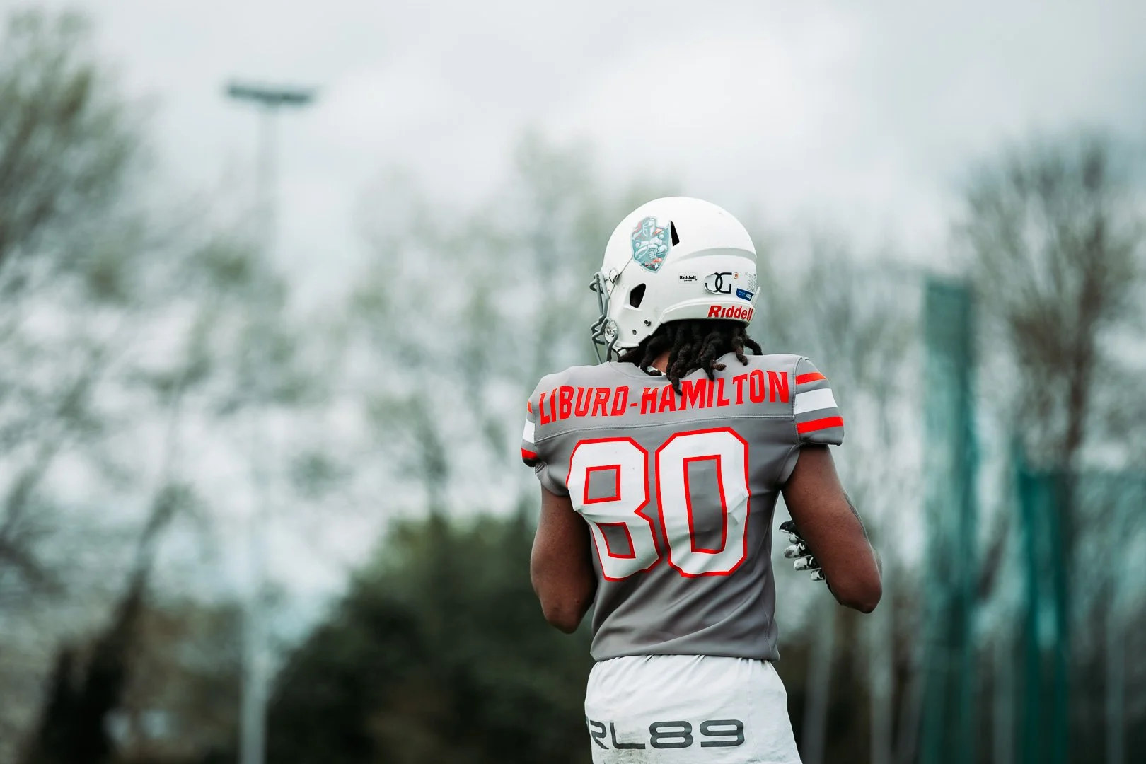 Football player from behind, wearing a gray jersey with the name Liburd-Hamilton and the number 30, white helmet with logos, on a cloudy outdoor field.