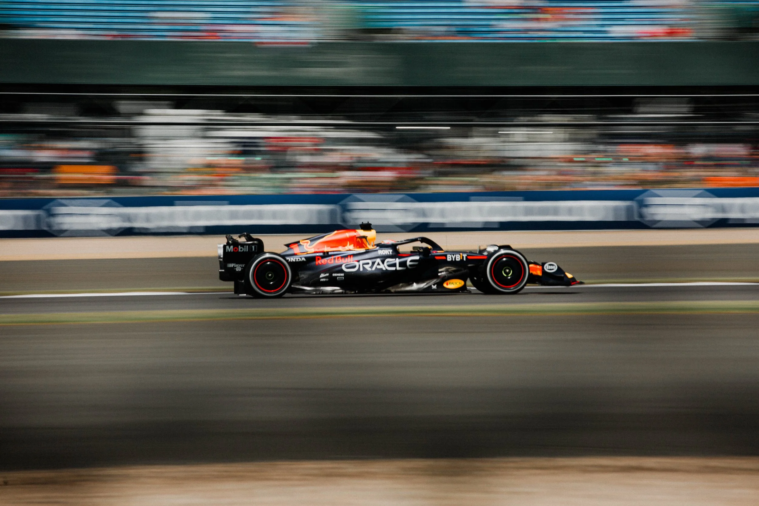 A Formula 1 race car speeding on a race track, with the background blurred to show motion. The car is black with orange and red accents, displaying sponsor logos including Oracle and Red Bull.