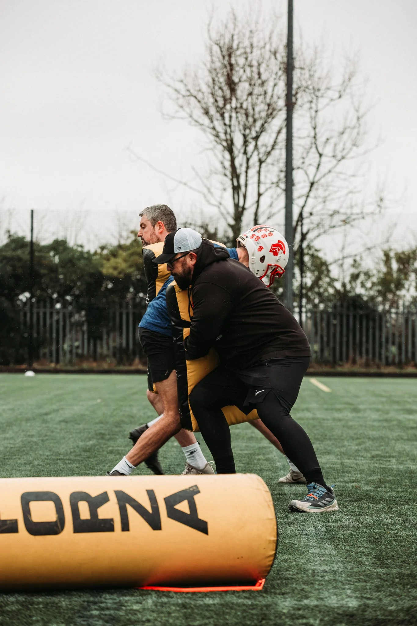 Two men practicing rugby on an outdoor field, with one man in a black jacket and leggings pushing against a padded dummy and the other man in athletic clothing, helmet, and pads. The scene is overcast with trees and a fence in the background.