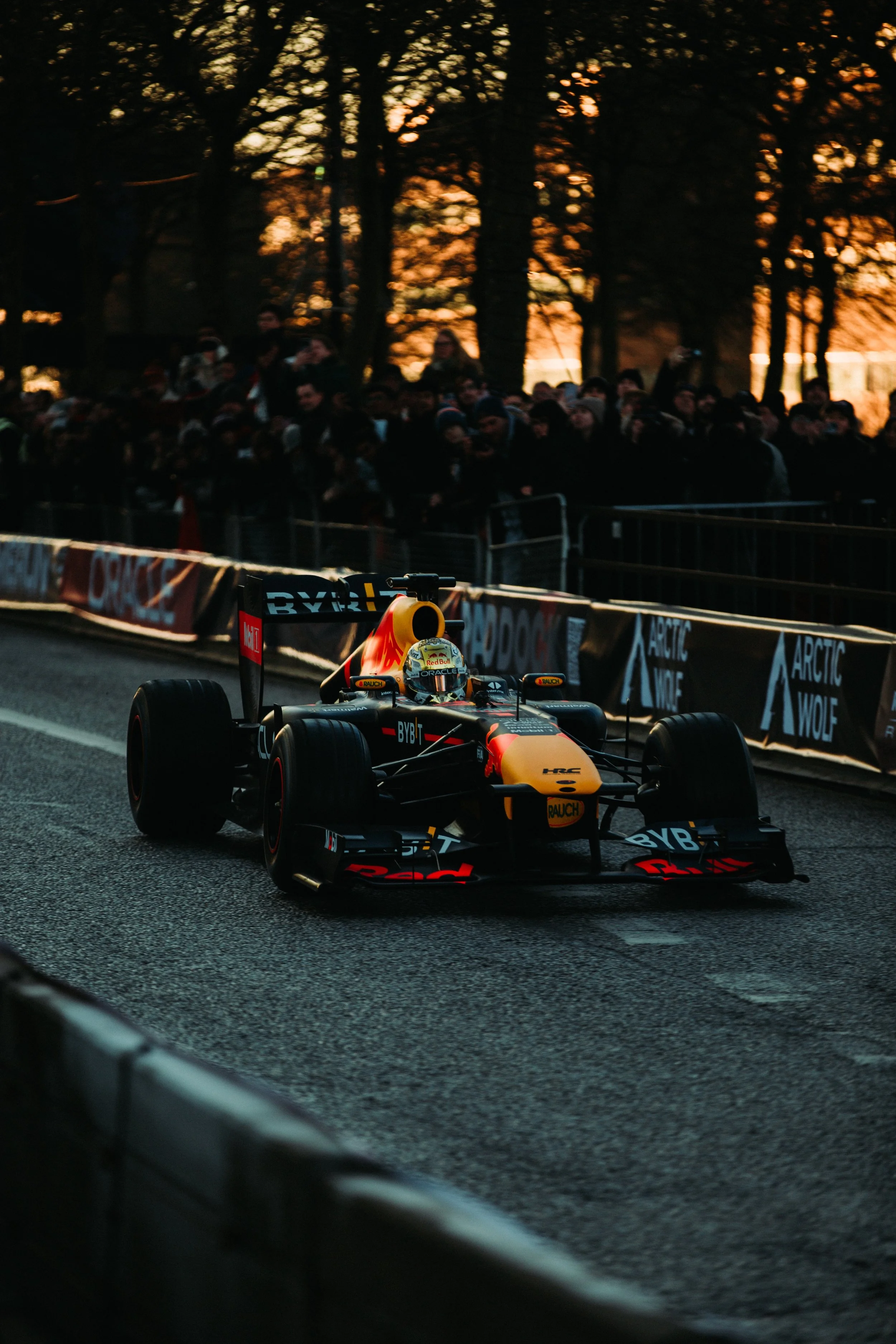 A Formula 1 race car driving on a track during sunset with spectators watching from behind safety barriers.