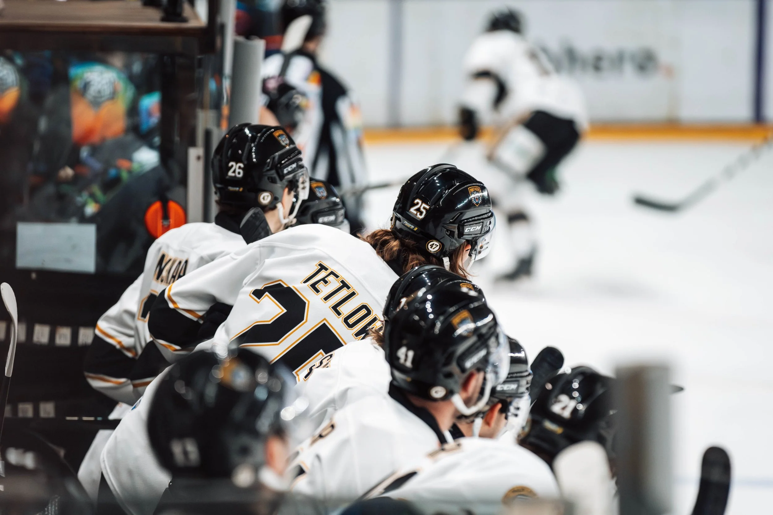 Hockey players in white jerseys and black helmets sitting on the bench during a game, with players skating in the background on the ice rink.