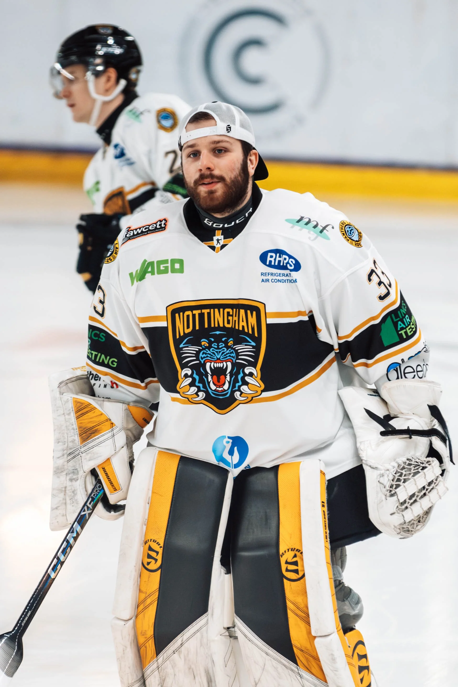 Ice hockey goalie in white Nottingham team jersey holding a hockey stick on ice rink with another player in white jersey in the background.