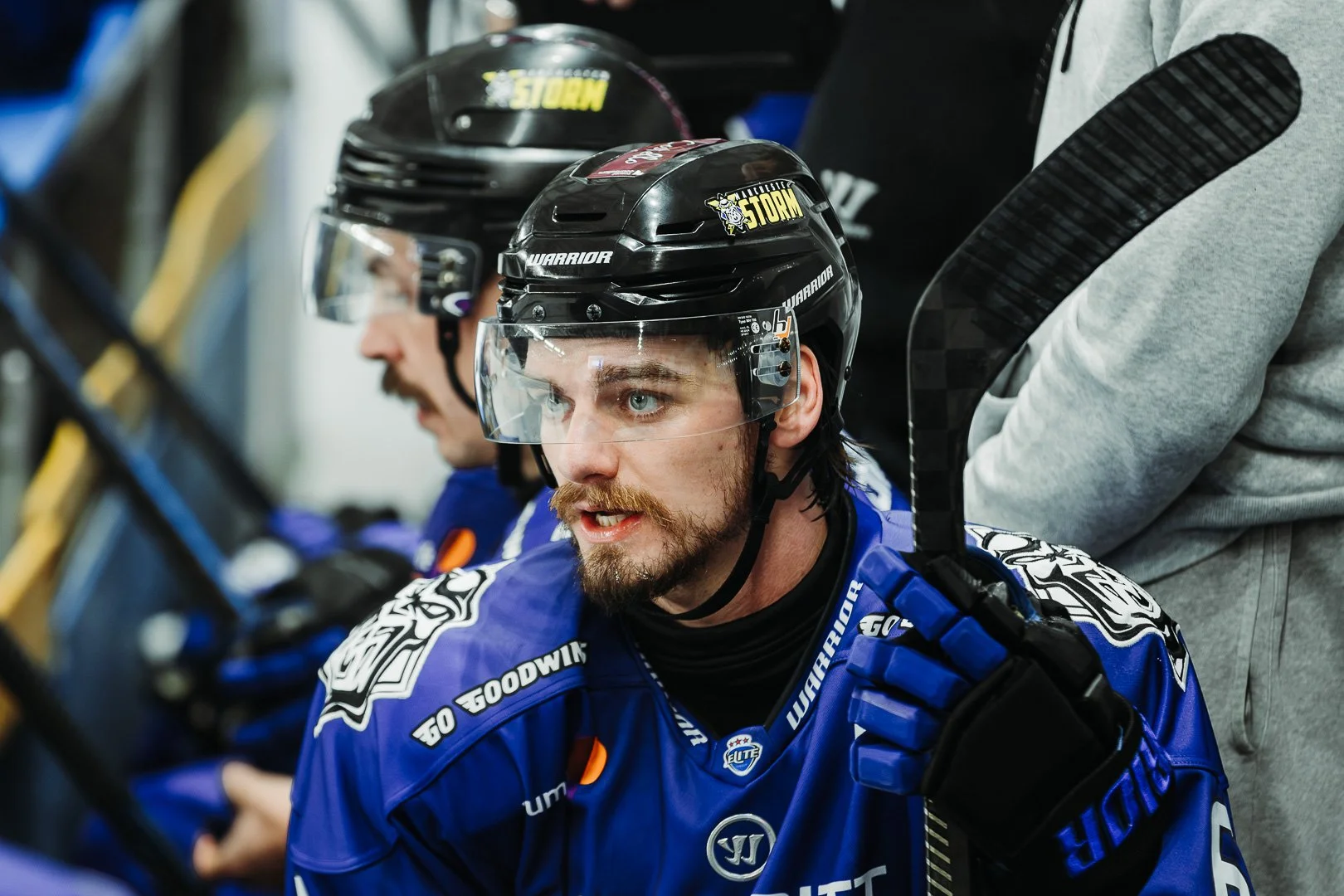 Hockey players wearing helmets and blue jerseys are sitting on the bench during a game, with one player looking alert and holding a hockey stick.