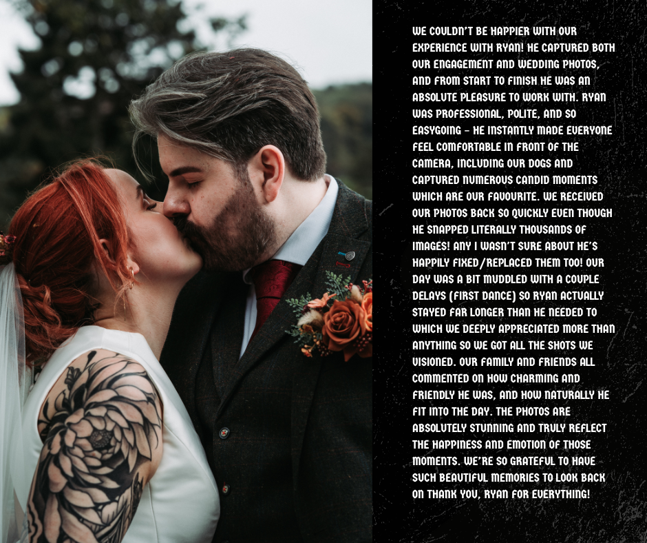 A couple is kissing outdoors during a wedding, with the bride having red hair and a tattoo on her arm, and the groom with brown hair and a beard. The groom is wearing a suit with a boutonniere.