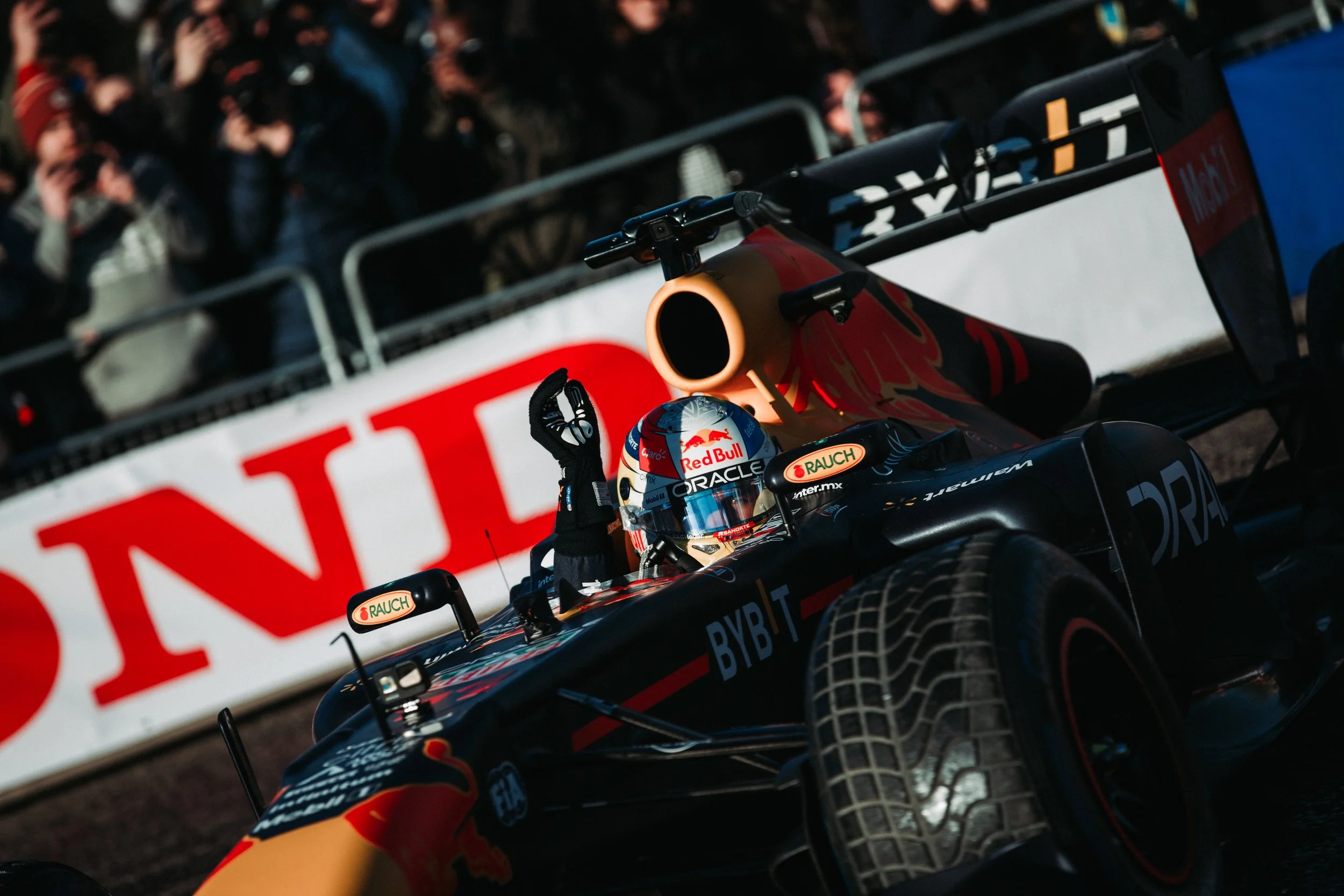 A Formula 1 race car with a driver giving a peace sign, on a track with a barrier and crowd in the background.