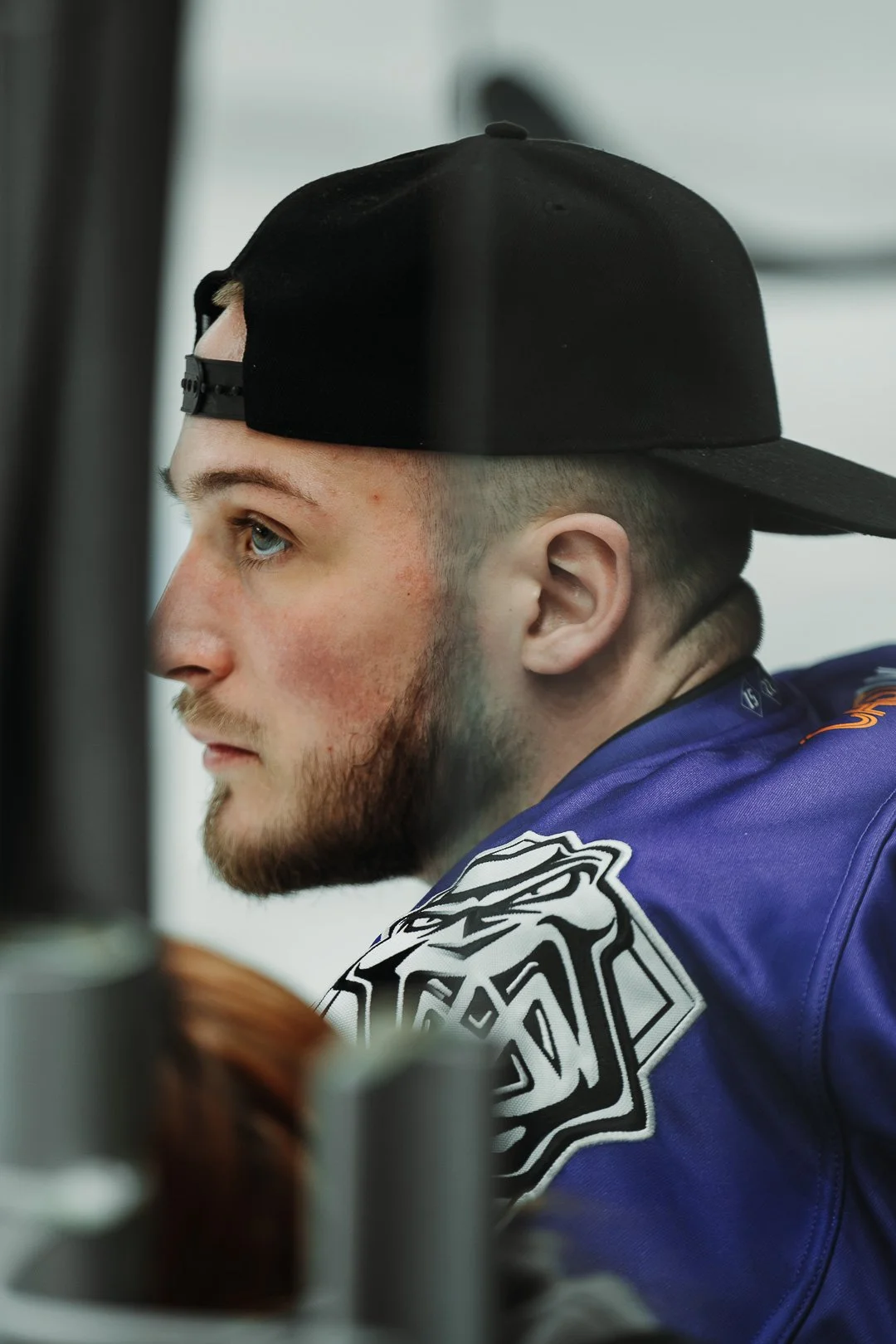 Close-up of a young man with a beard, blue eyes, wearing a black cap backwards and a blue sports jersey with a white logo, sitting near a window.