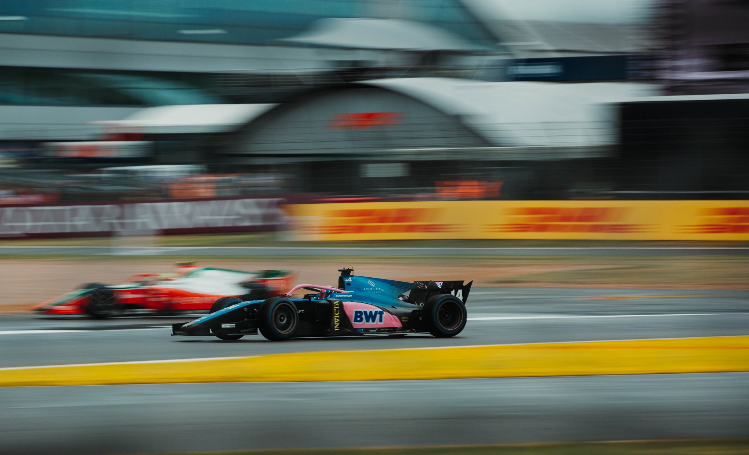 A Formula 2 race car speeds on a racetrack in front of a blurred grandstand and advertising banners.