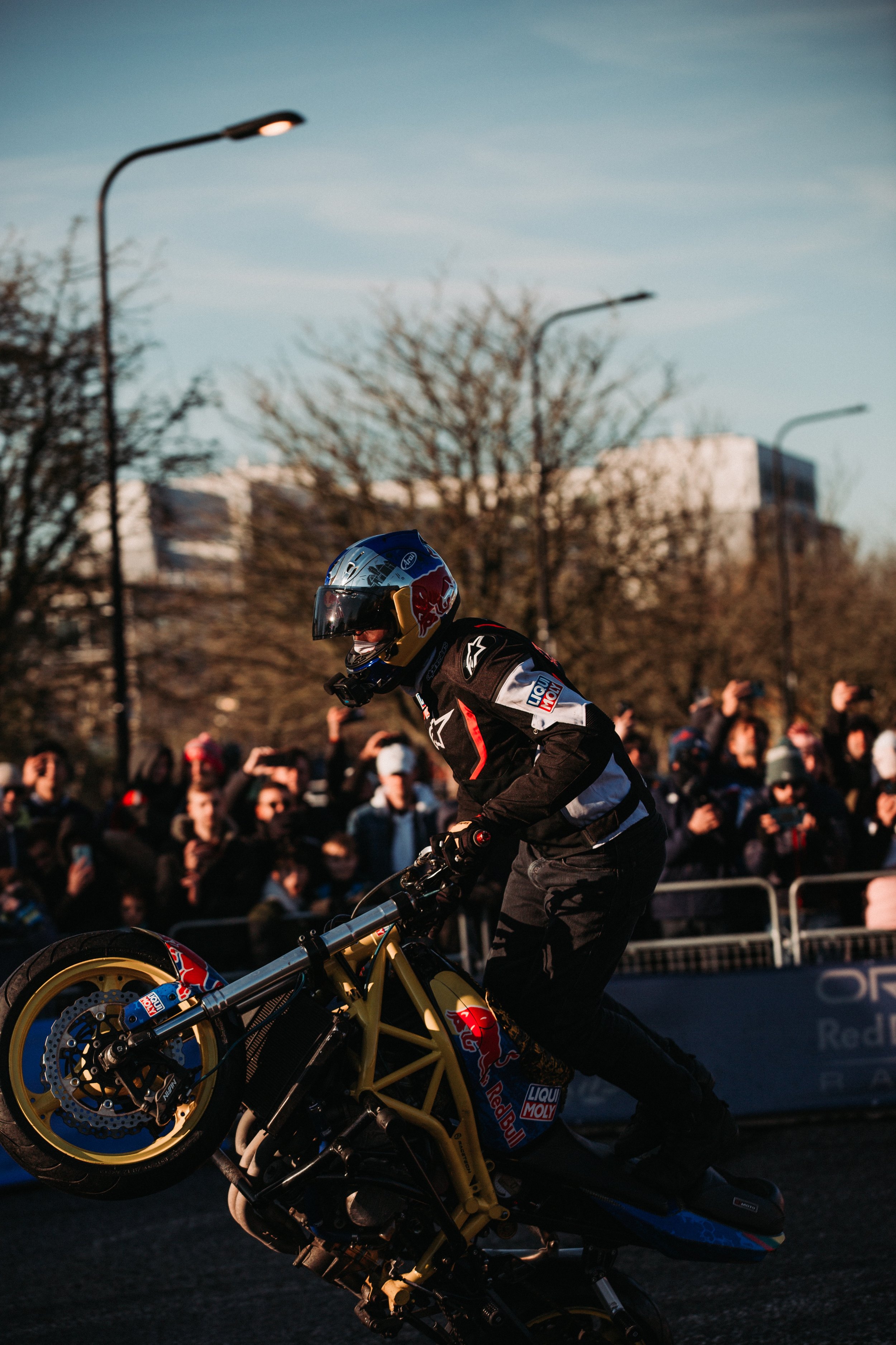 Motorcycle stunt rider performing a wheelie in front of a crowd during a daytime event.