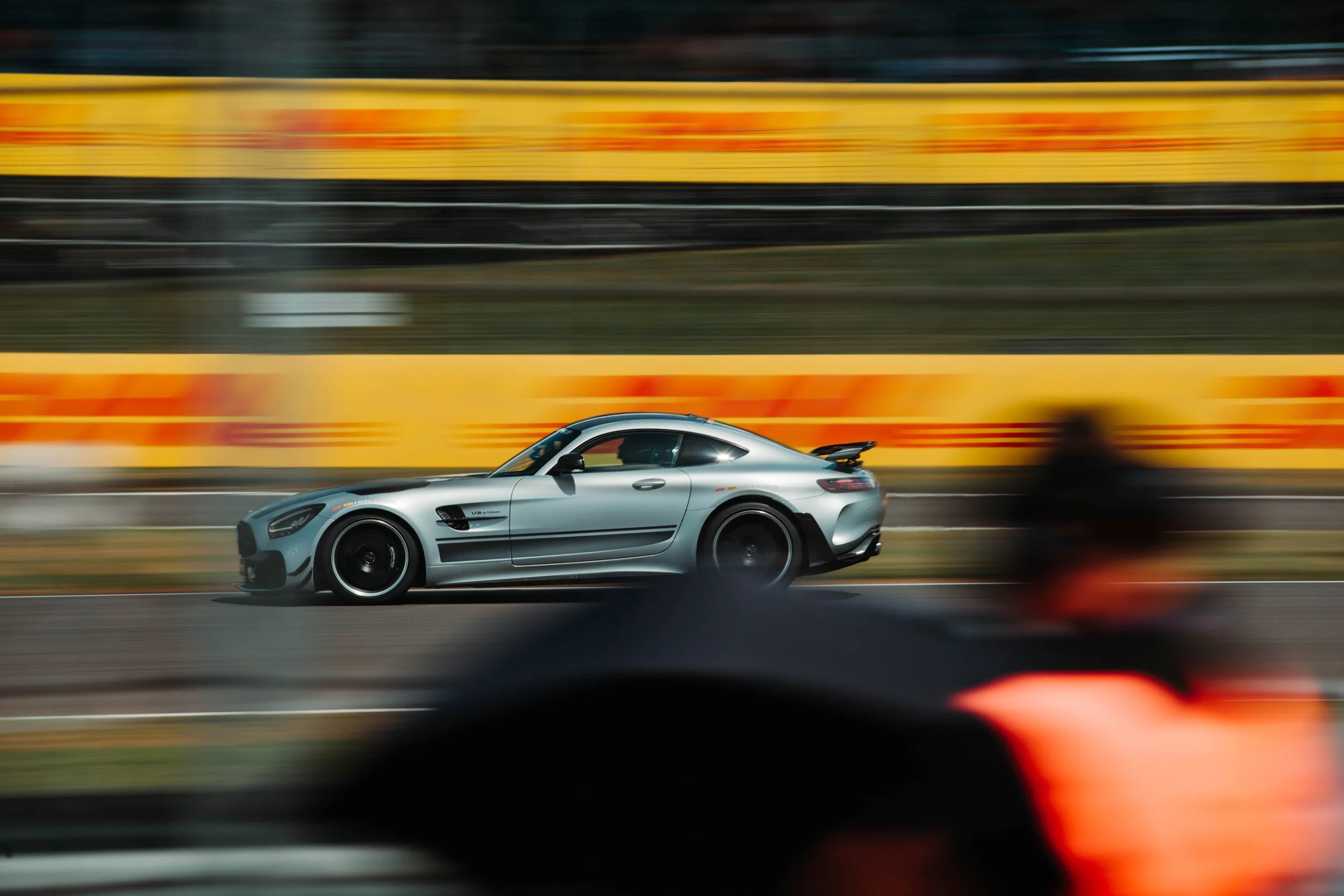 A silver Mercedes-Benz sports car speeding on a race track with a blurred background of yellow and orange barriers and a blurry foreground of another vehicle.