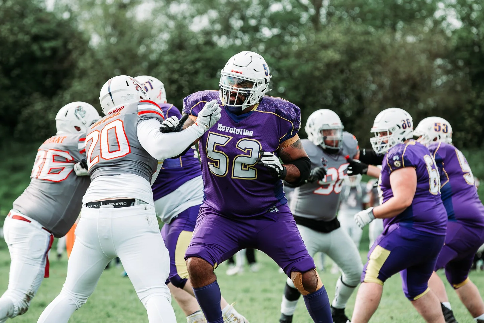 American football players in purple and gray uniforms engaging during a game on a grass field with trees in the background.