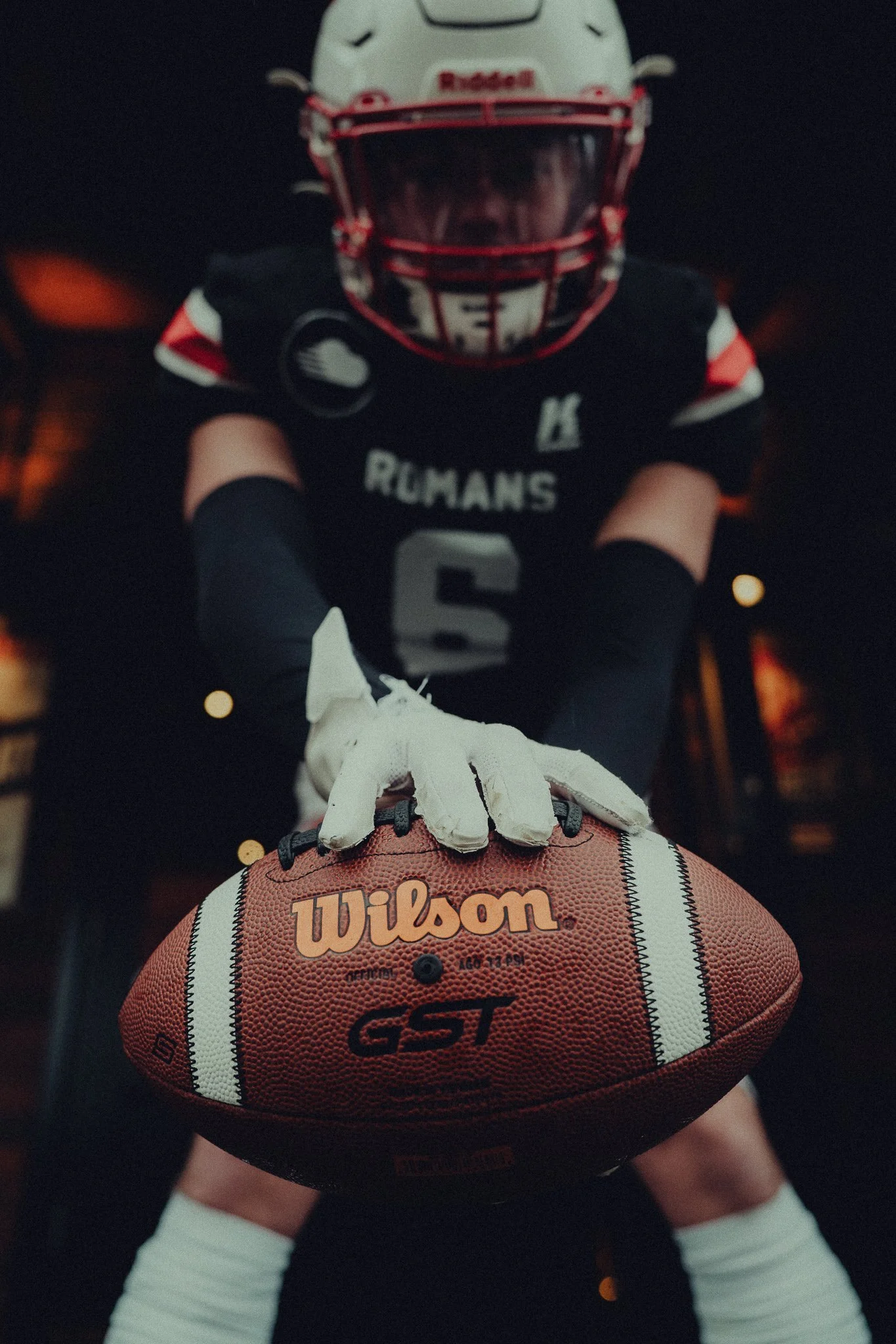 A football player in full gear leaning forward with one hand on a Wilson football, wearing a helmet and jersey, in a dark indoor setting.