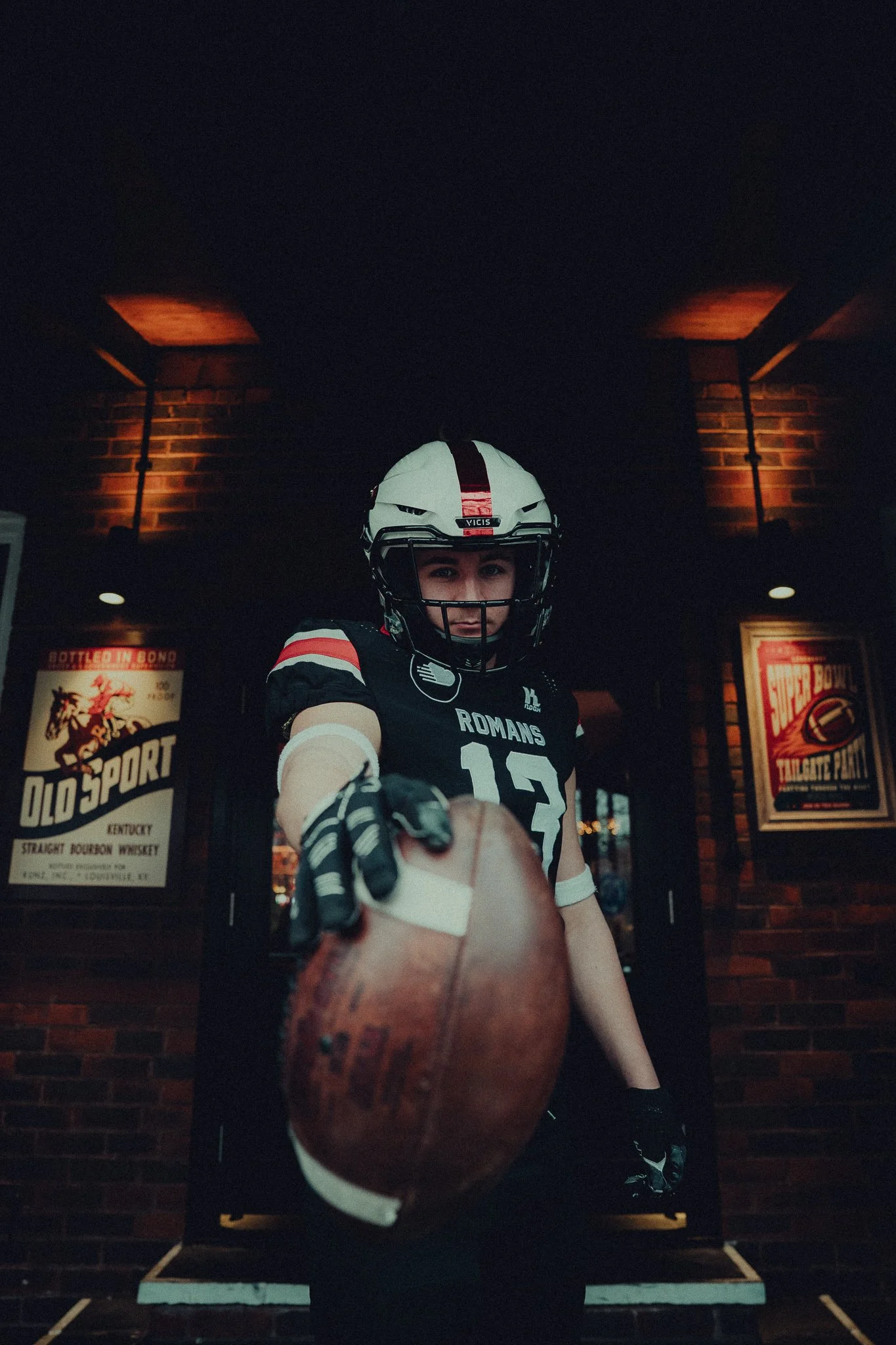 A woman in football gear extending her hand with a football towards the camera, in an indoor setting with brick walls and vintage signs.