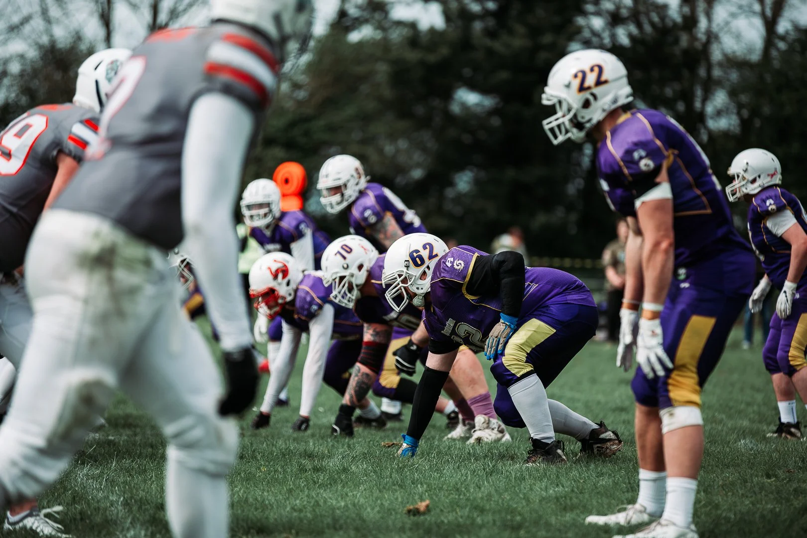 American football players in purple and white uniforms lined up on the field, preparing for a play. The players are in a crouched stance, ready to engage, with a grassy field and trees in the background.