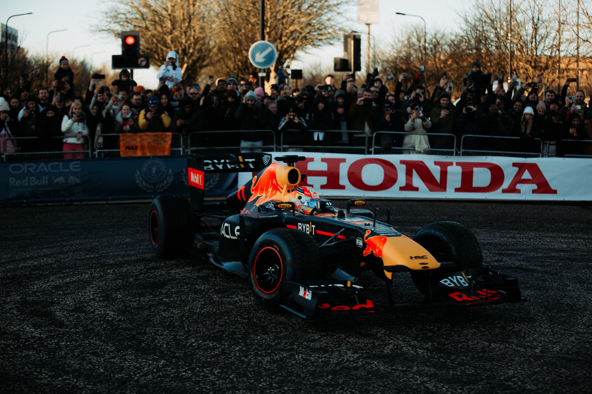 A black and orange Formula 1 car racing on a dark track with a crowd of spectators behind safety barriers, and banners with logos of Honda and Oracle Red Bull Racing.