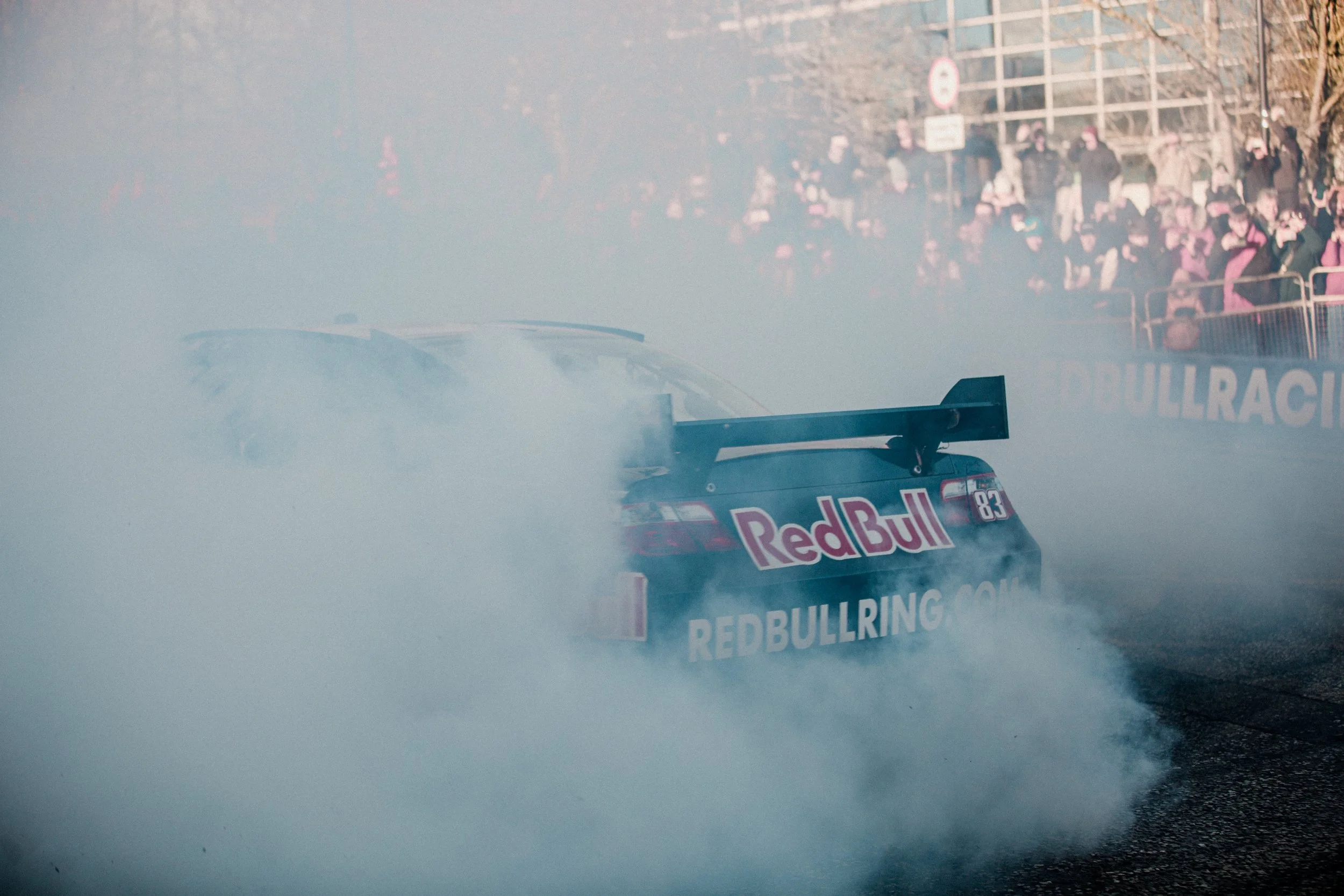 A race car with Red Bull branding performing a burnout, creating a smoke cloud on the track during an event with spectators and barriers in the background.