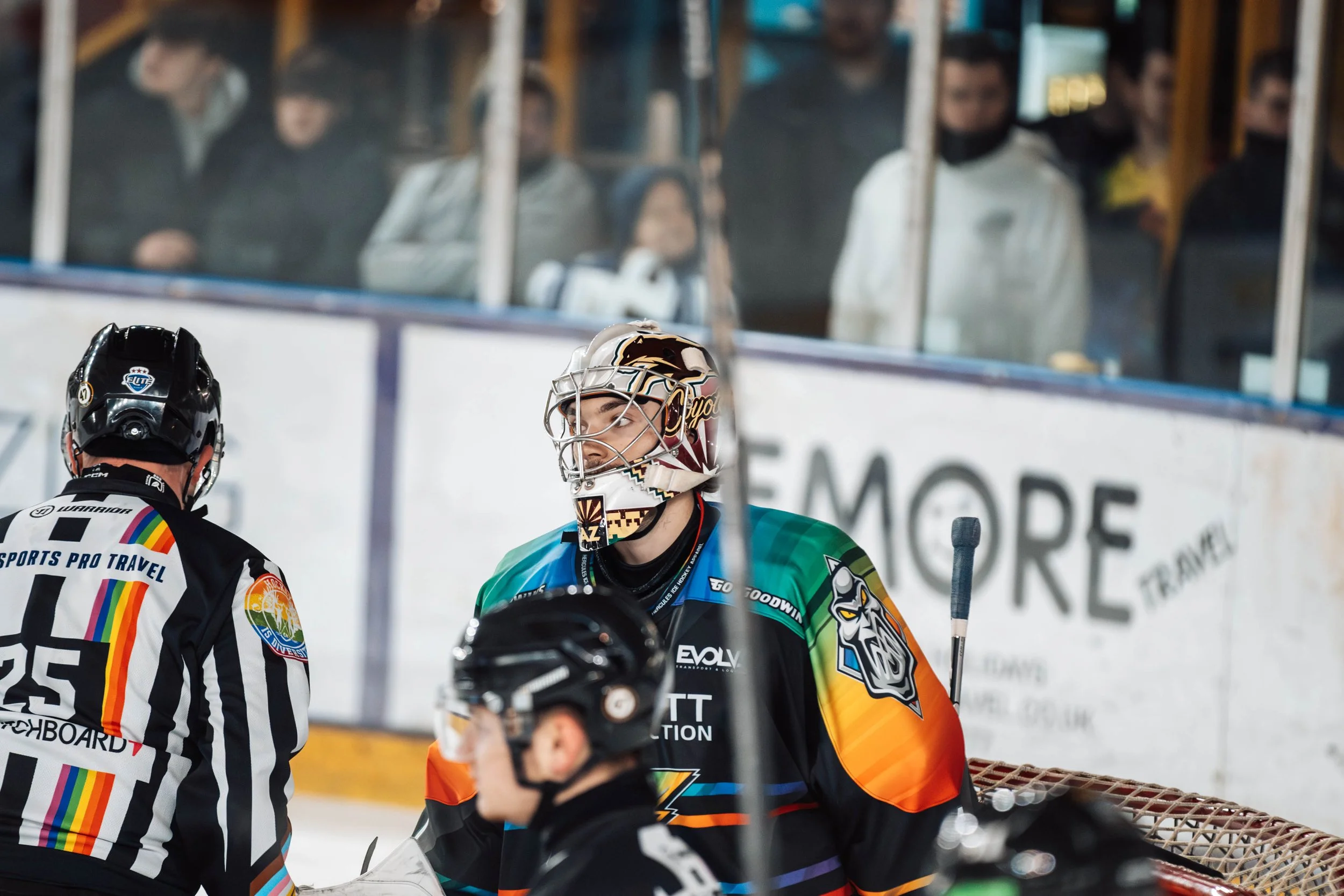 Hockey players on ice rink, one in colorful jersey, referee in striped uniform, and spectators watching behind glass