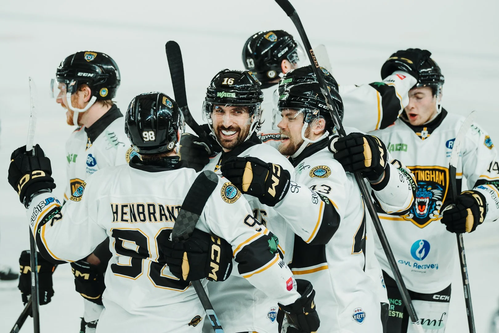 Ice hockey players celebrating on the ice, wearing white jerseys and helmets, some with their arms around each other, showing excitement and camaraderie.