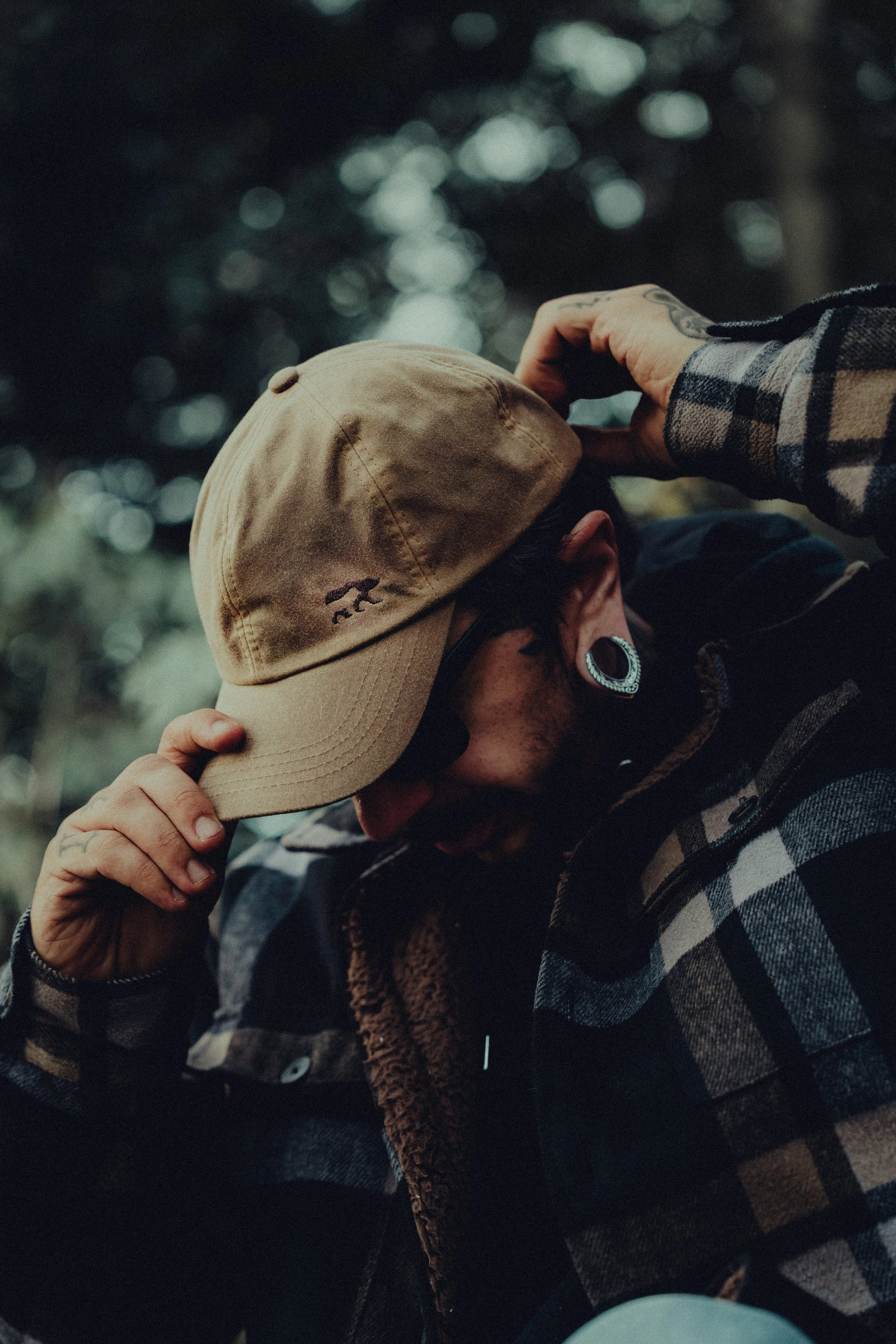 Person adjusting a tan cap outdoors, wearing a plaid jacket and earrings.