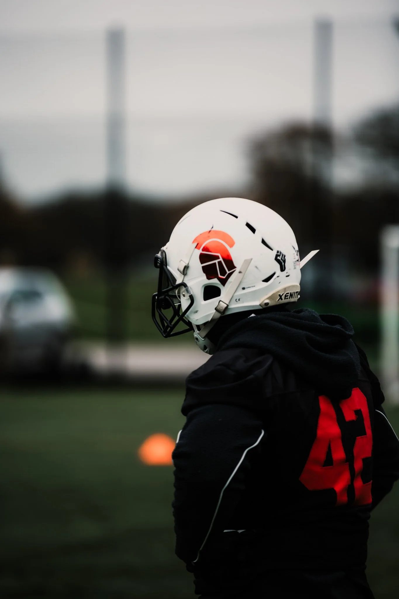 A football player wearing a black hoodie and a white helmet with a Spartan logo on it, standing on a field with an orange cone in the background.