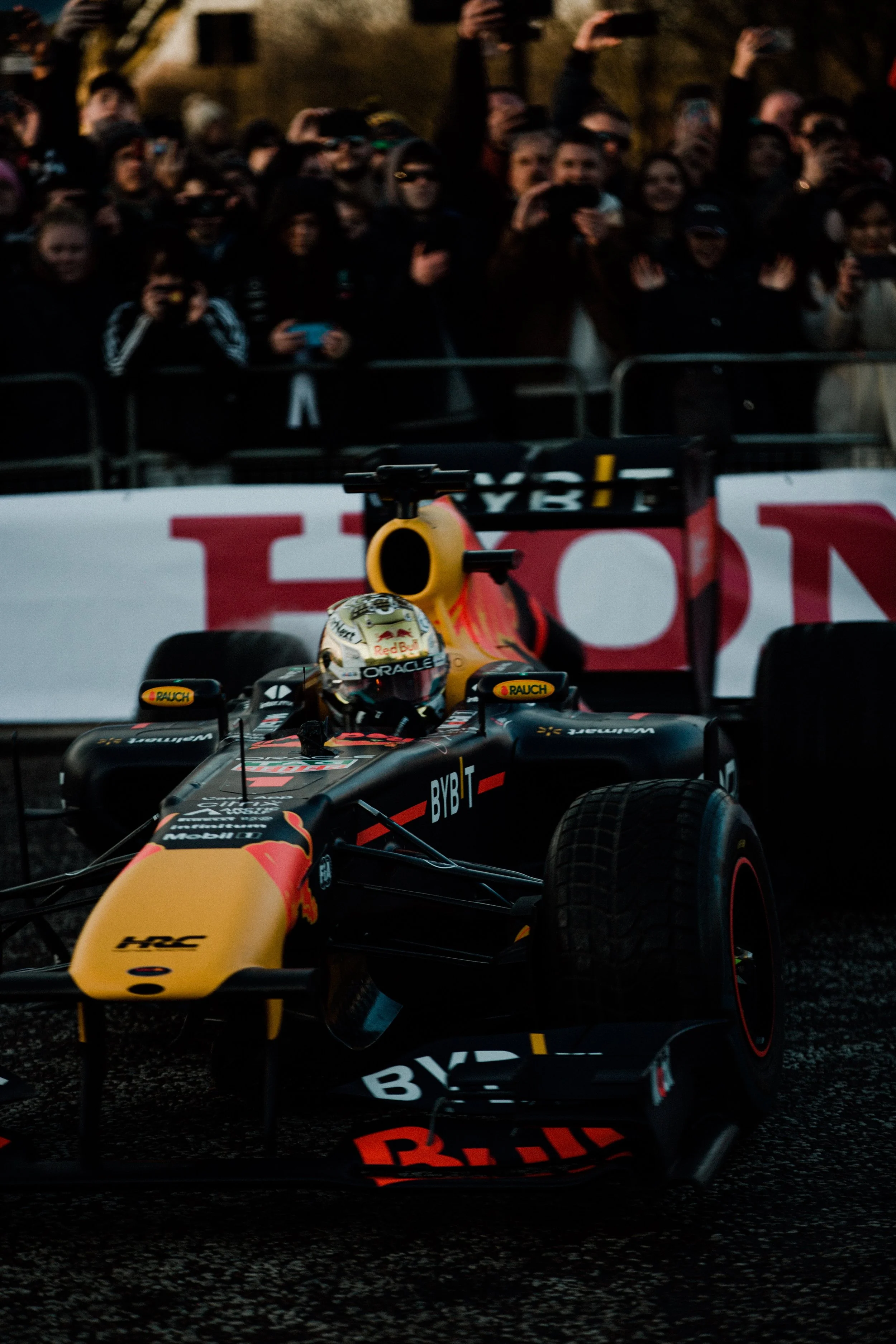 A Formula 1 race car with black and yellow livery on track, surrounded by spectators behind barriers, during evening or night.