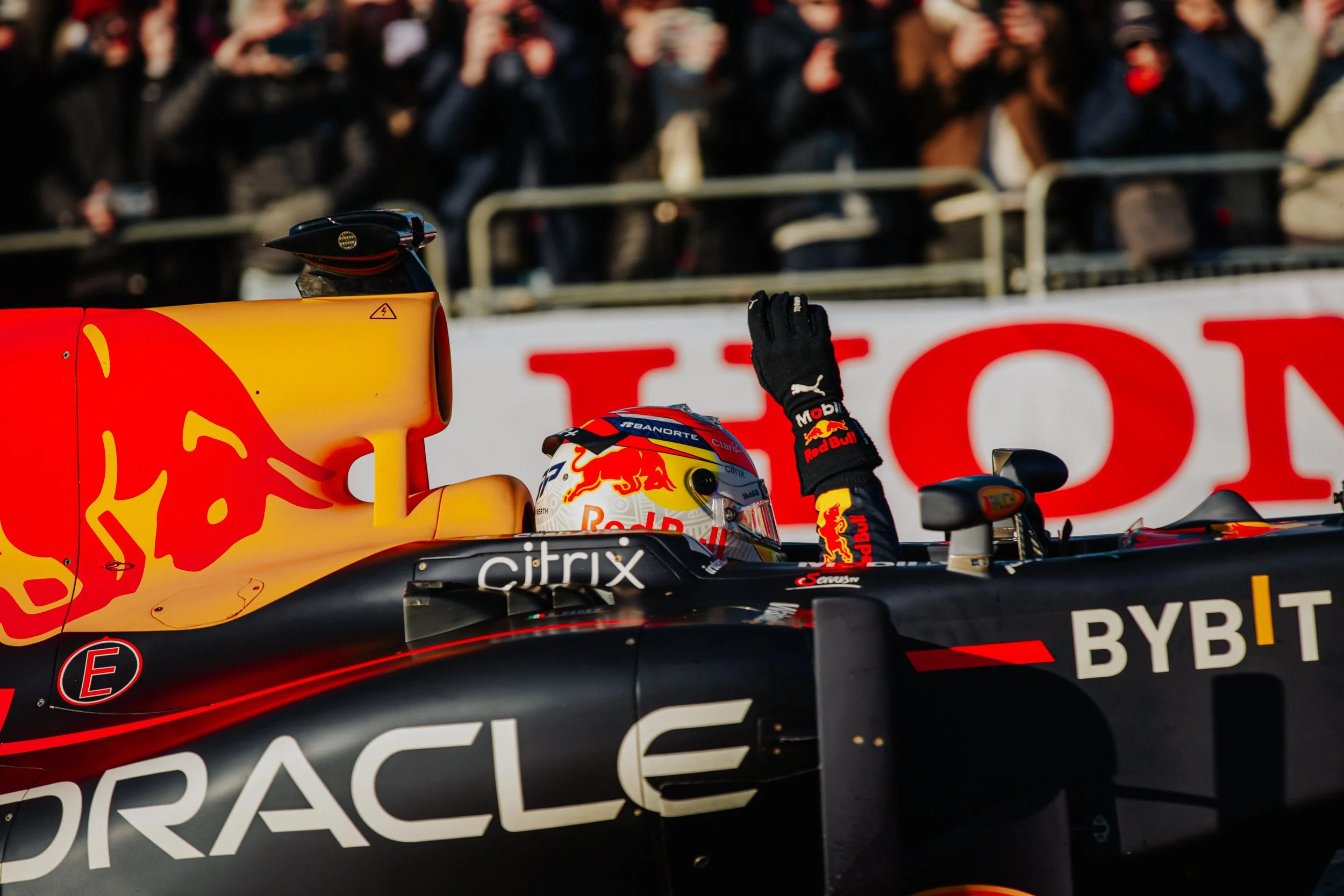 Race car driver in a Red Bull racing car celebrating, waving to the crowd with a gesture, wearing a helmet with Red Bull branding, behind safety barriers with spectators in the background.