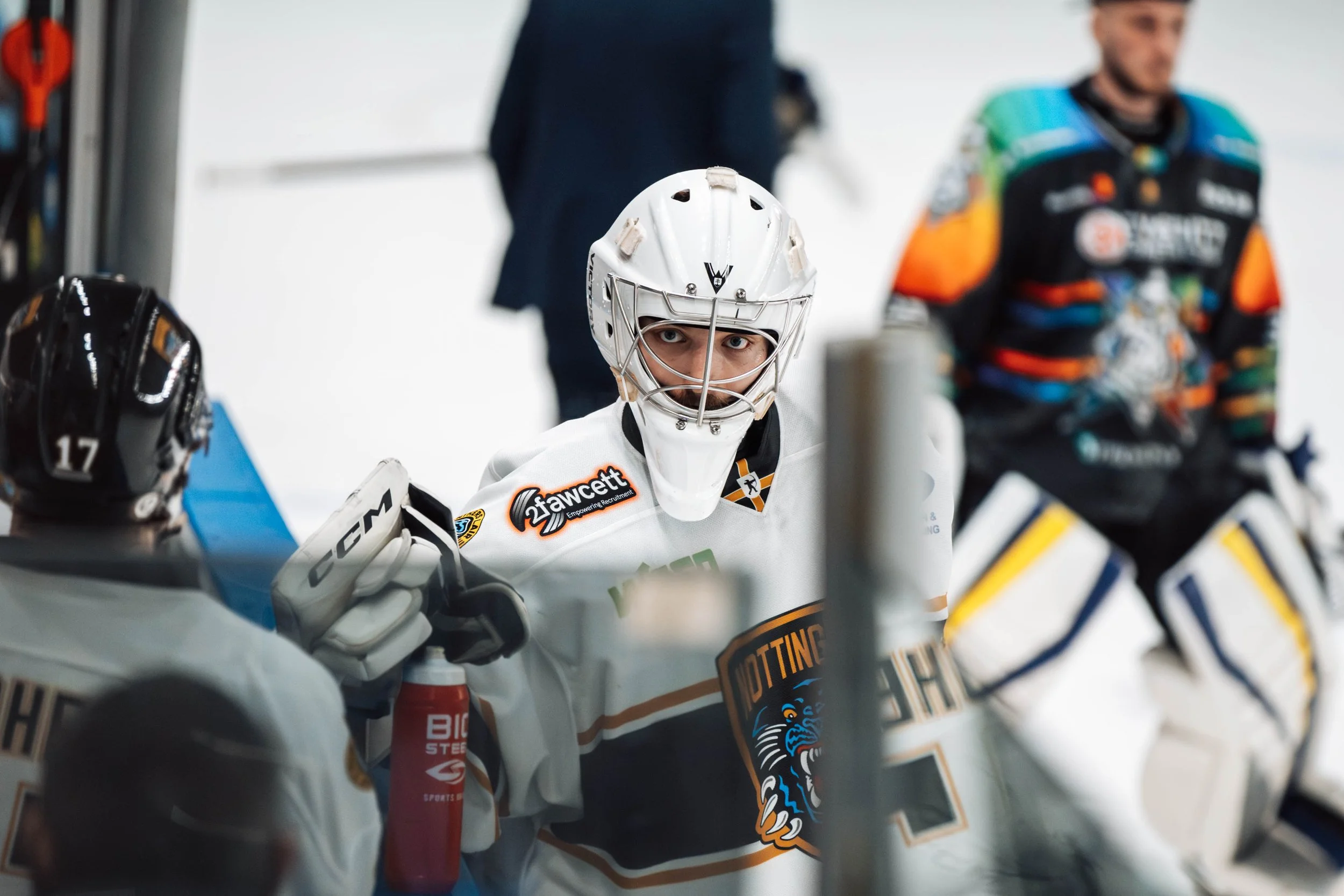 An ice hockey player wearing a white jersey and helmet, sitting on the bench with a focused expression, surrounded by teammates.