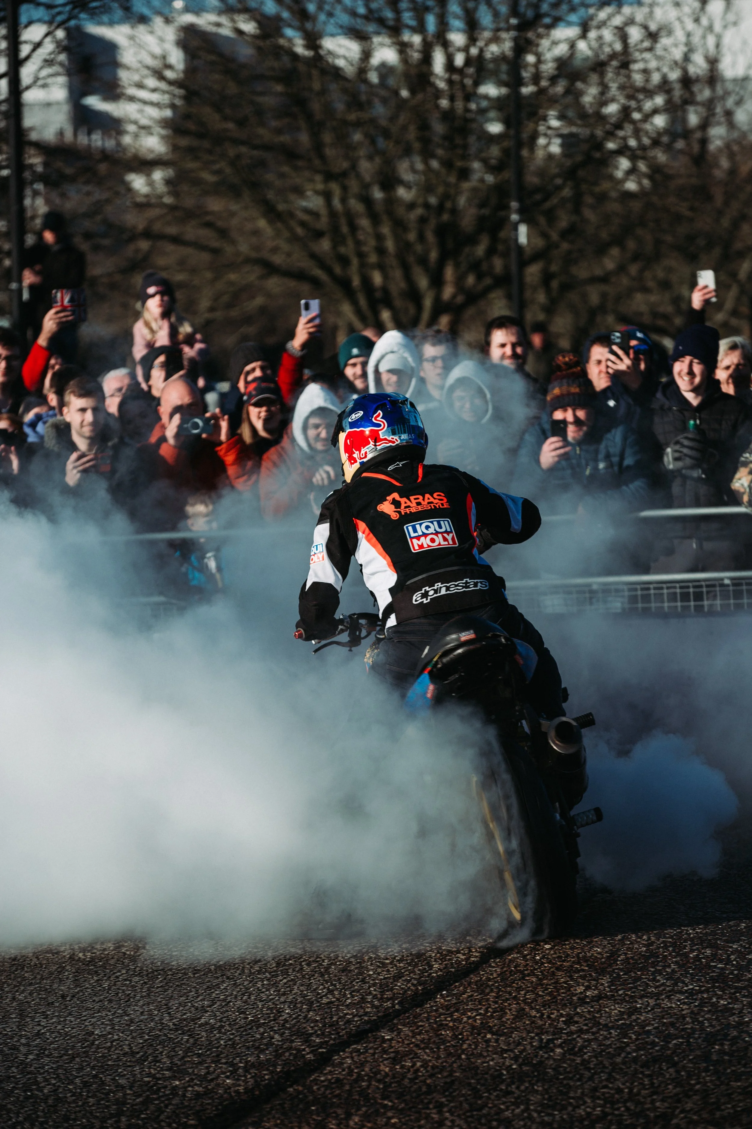 Motorcycle stunt rider performing burnout in front of a crowd of spectators taking photos and videos, with smoke from the tires visible.